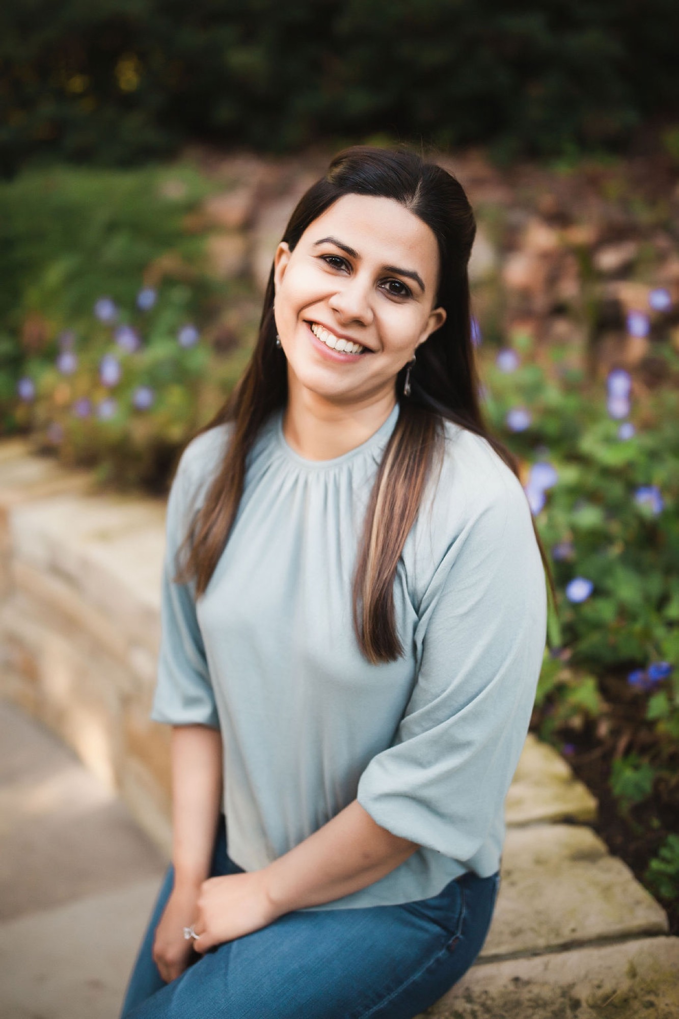 Woman kneeling on a stone path smiles warmly at the camera with purple flowers blooming softly behind her in dappled garden light