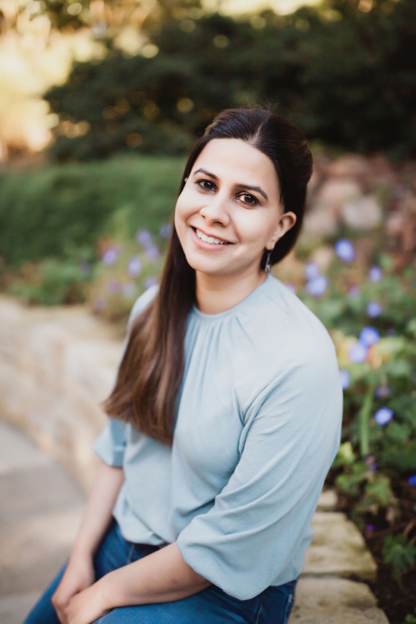 Woman in soft blue shirt smiles warmly over her shoulder, seated among purple flowers in a lush garden setting with golden afternoon light