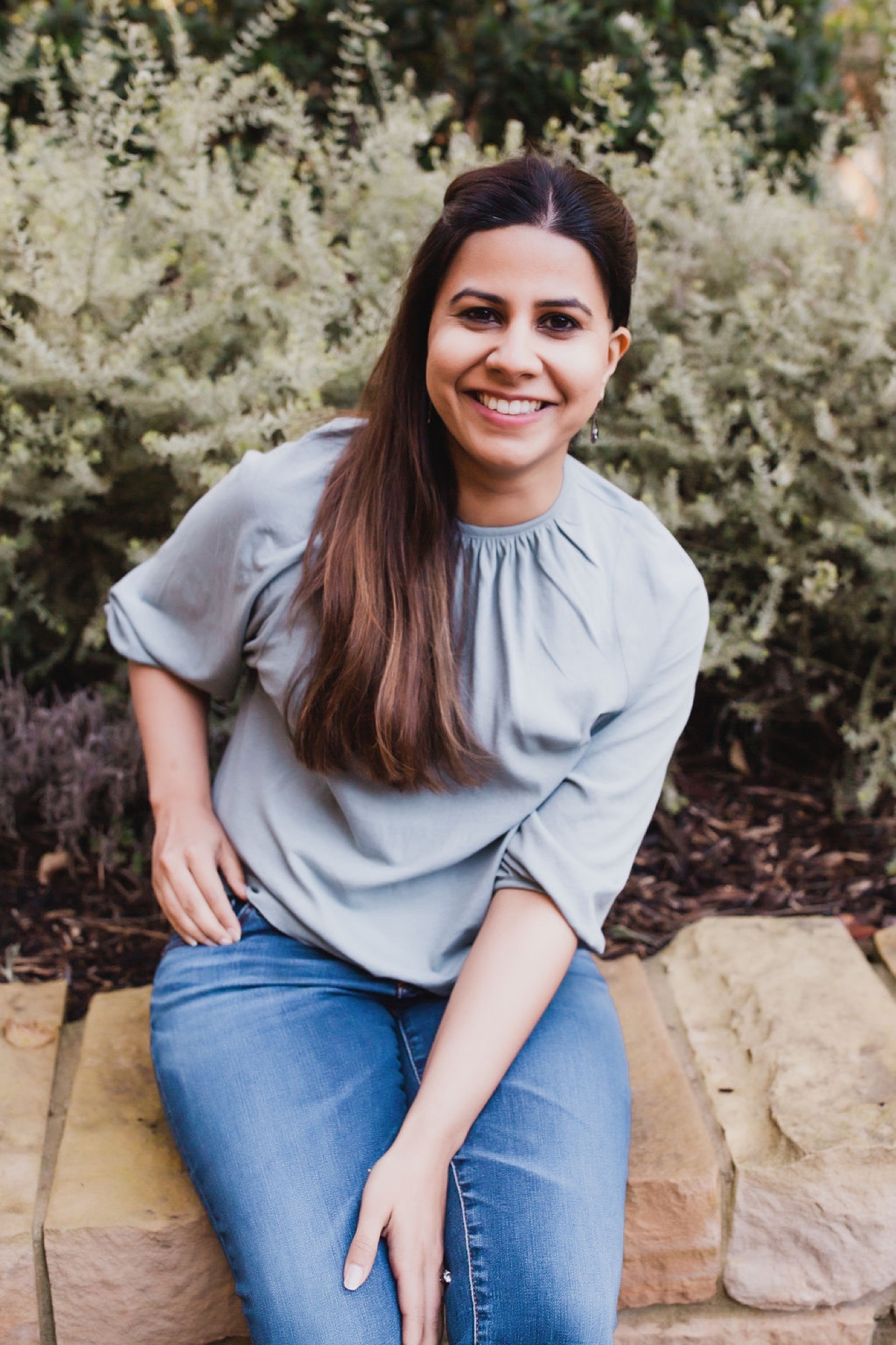 Woman in light gray shirt and jeans smiles warmly while seated on stone ledge, backlit by flowering shrubs