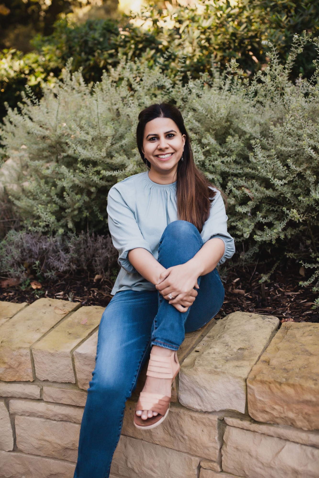 Woman in light blue shirt and distressed jeans sits on weathered stone blocks, smiling warmly with silvery-green foliage glowing softly behind her