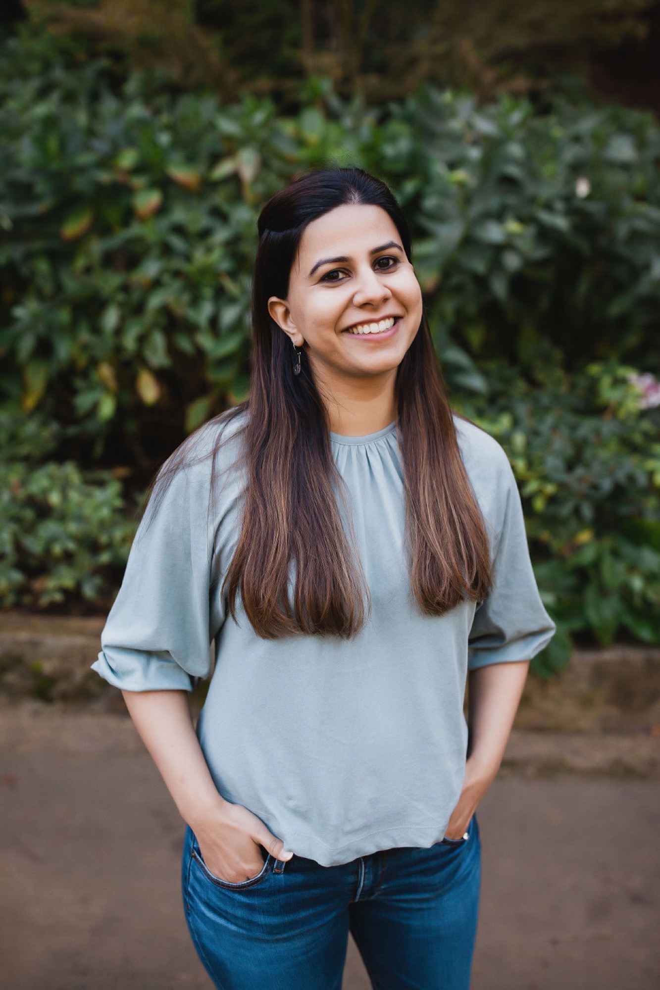 Woman with long dark hair smiles warmly at camera, wearing light blue shirt and jeans against lush green foliage backdrop