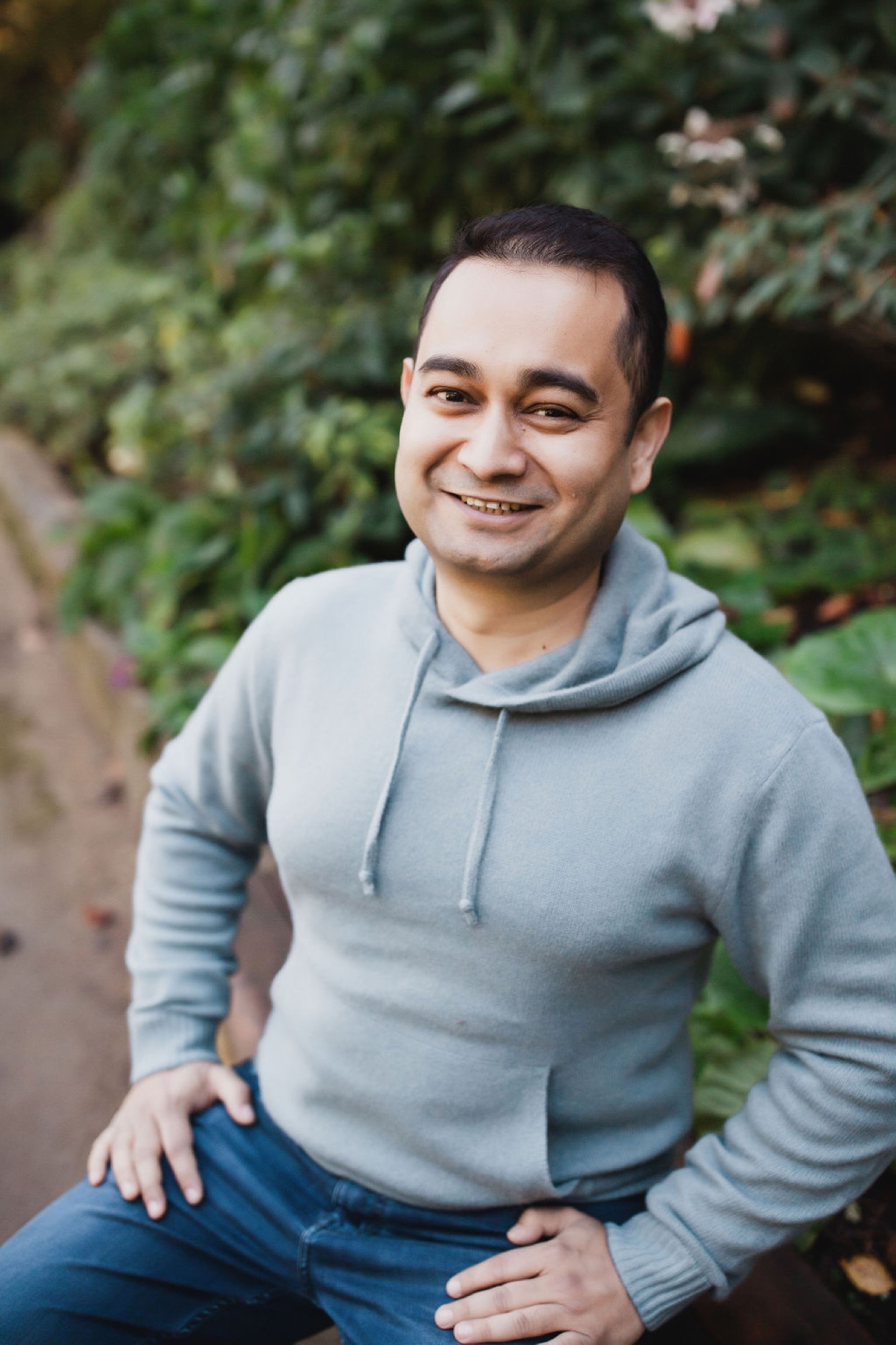 Man in light gray hoodie and jeans smiles warmly at camera, seated against lush green ivy-covered backdrop with soft natural light
