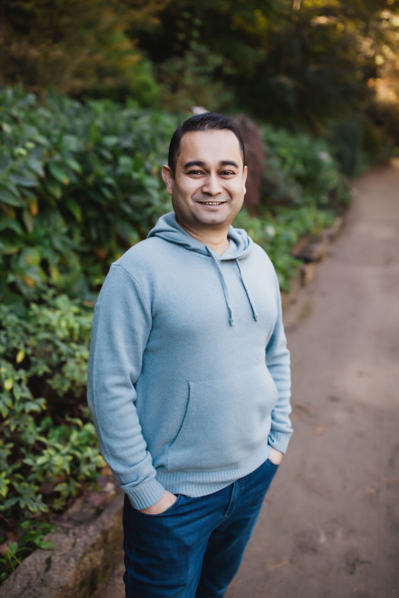 Man in light blue hoodie smiles warmly at camera while standing on a tree-lined garden path with soft natural light filtering through foliage