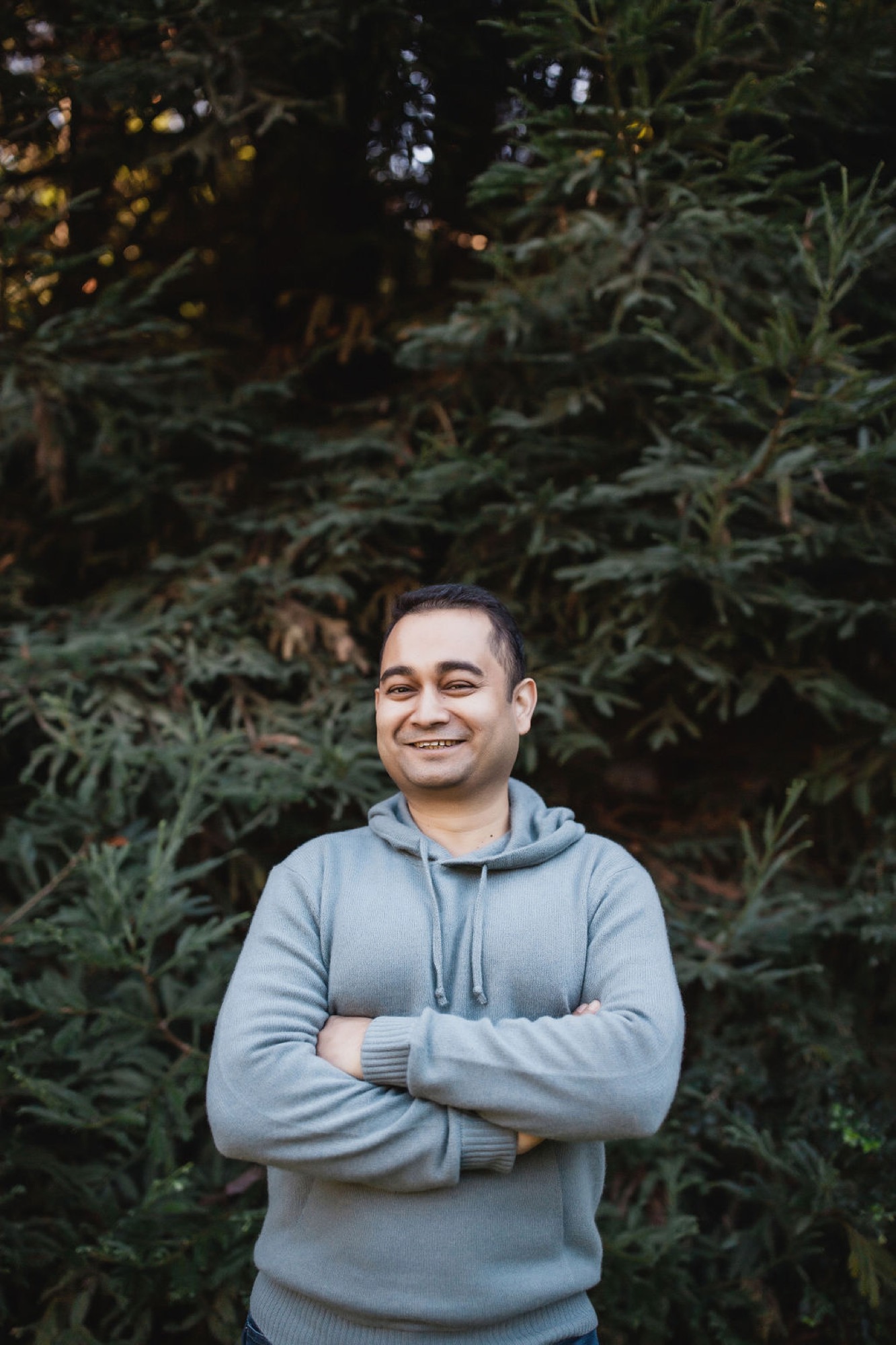 Man in light blue hoodie stands confidently with crossed arms before dense evergreen foliage, genuine smile and direct gaze