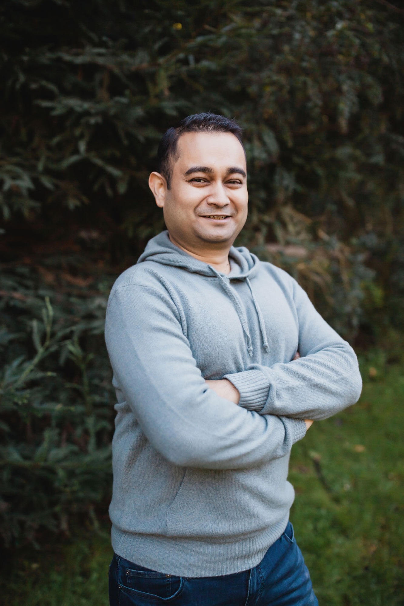 Man in light gray hoodie stands with arms crossed, smiling warmly at camera against dark evergreen foliage backdrop
