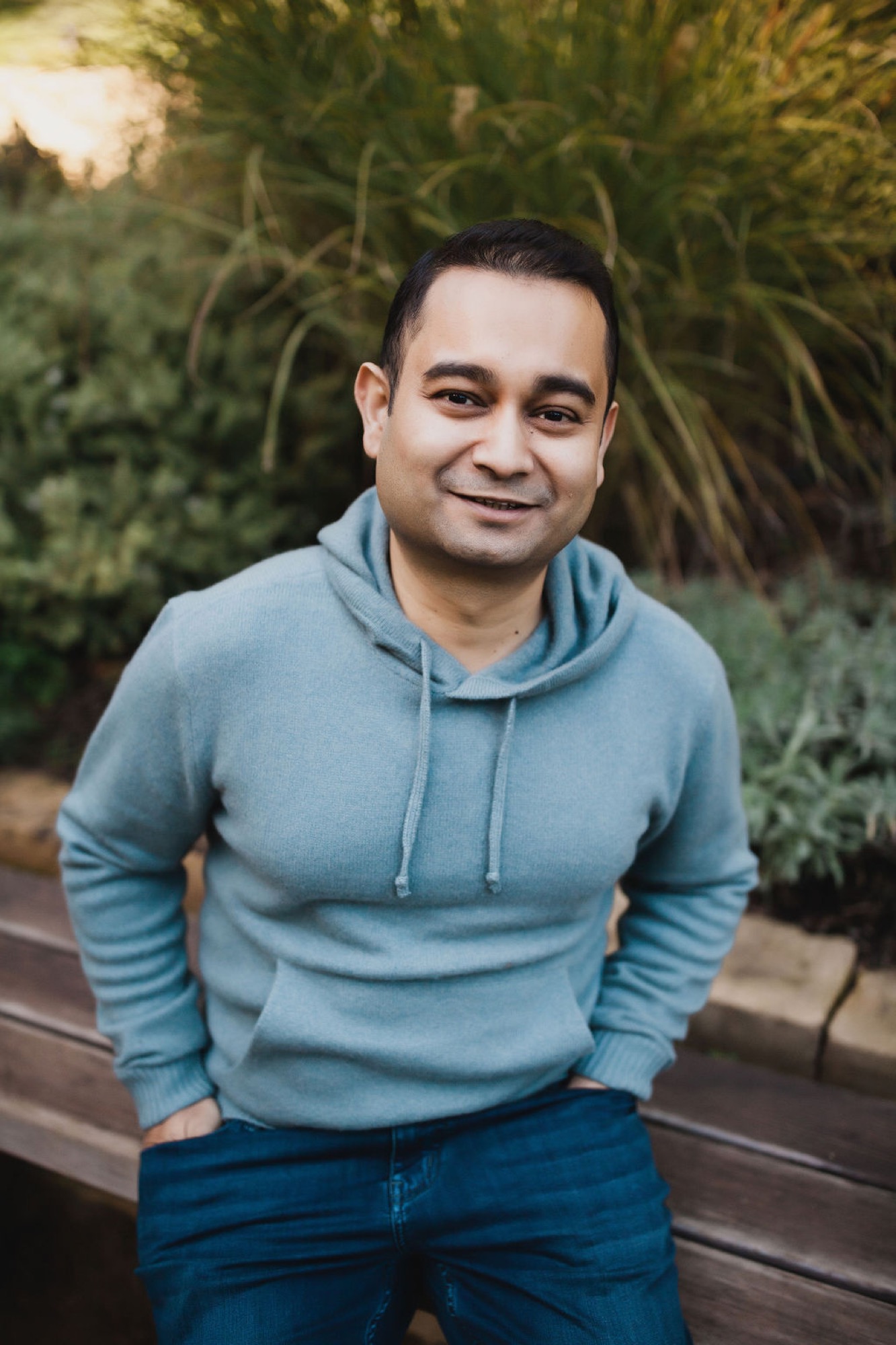 Man in blue hoodie smiles warmly at camera while standing against lush green foliage backdrop
