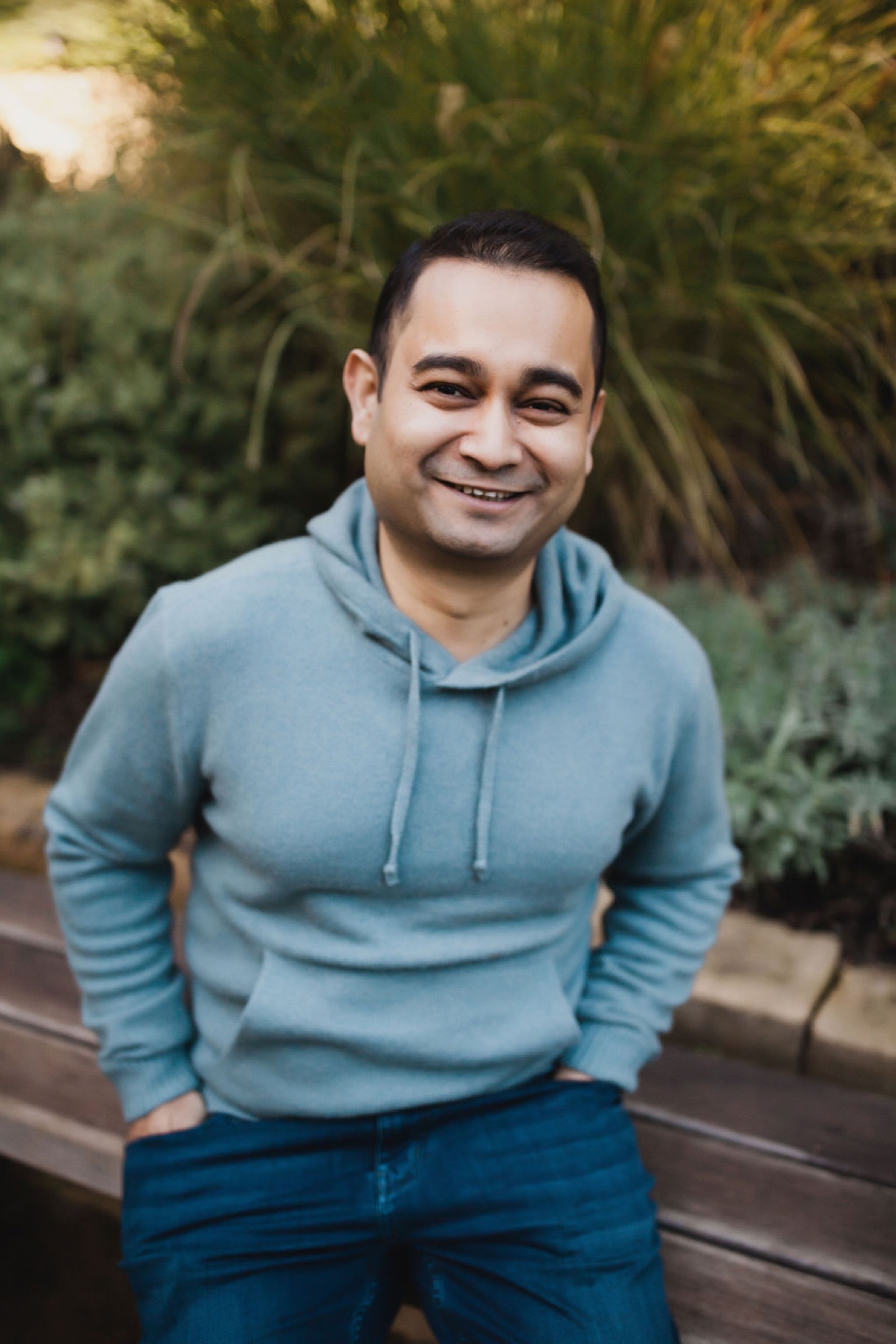 Man in light blue hoodie smiles warmly at camera with lush green foliage creating soft natural backdrop behind him
