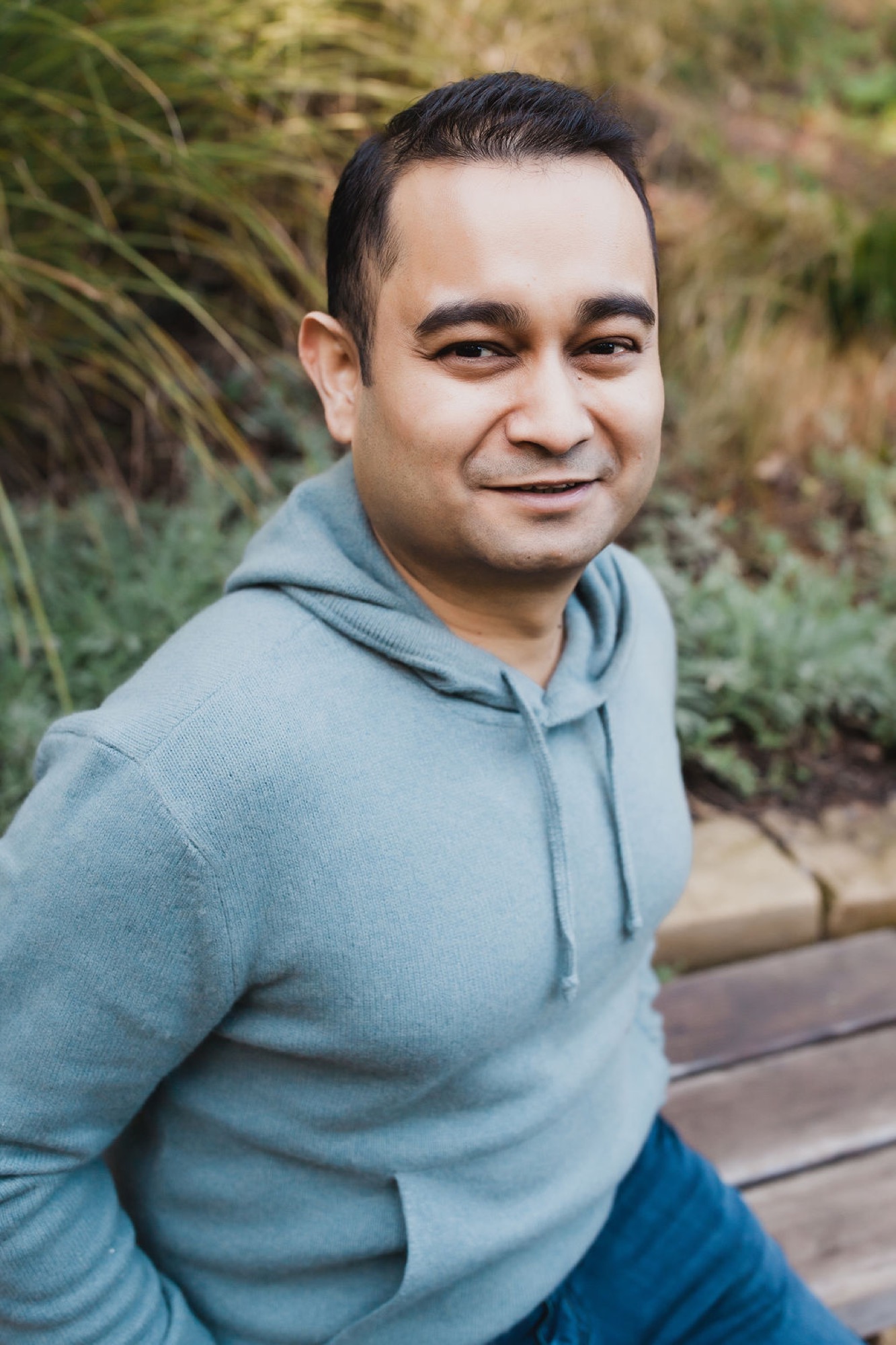 Man in light blue hoodie smiles warmly over his shoulder against blurred green foliage backdrop