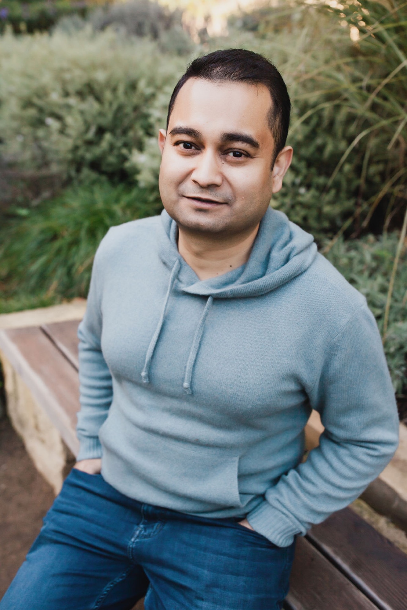 Man in light blue hoodie and jeans sits thoughtfully on a wooden bench, surrounded by soft green foliage with warm natural light