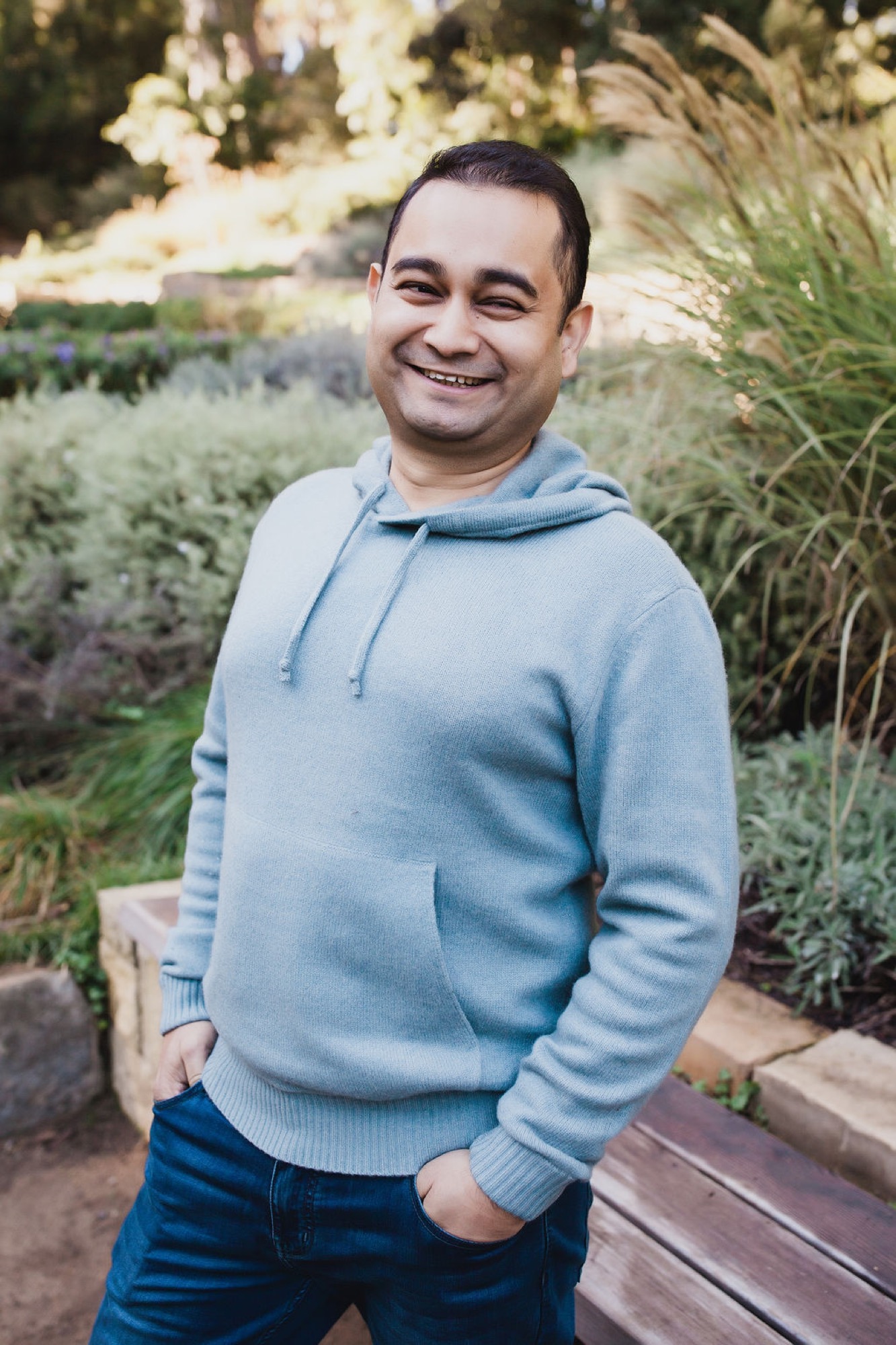 Man in light blue hoodie smiles warmly at camera while standing in sunlit garden with blurred foliage background