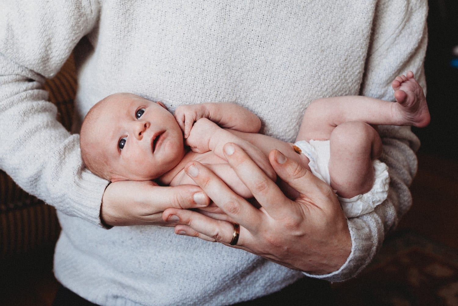 Dad in a cream sweater cradles his wide-eyed newborn in his arms
