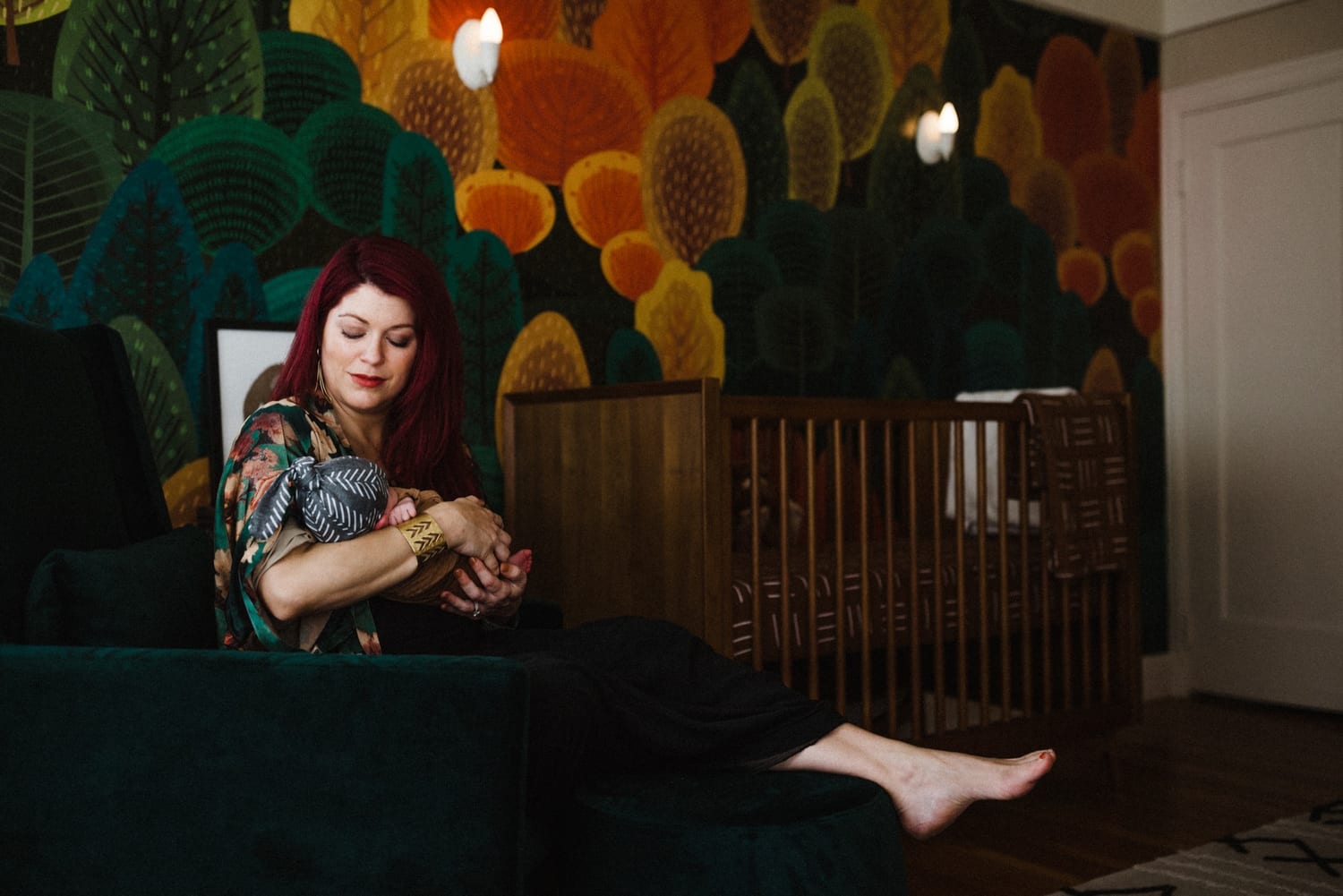 Mom in a floral kimono snuggles her newborn beside the crib and forest mural, in-home newborn photography San Francisco
