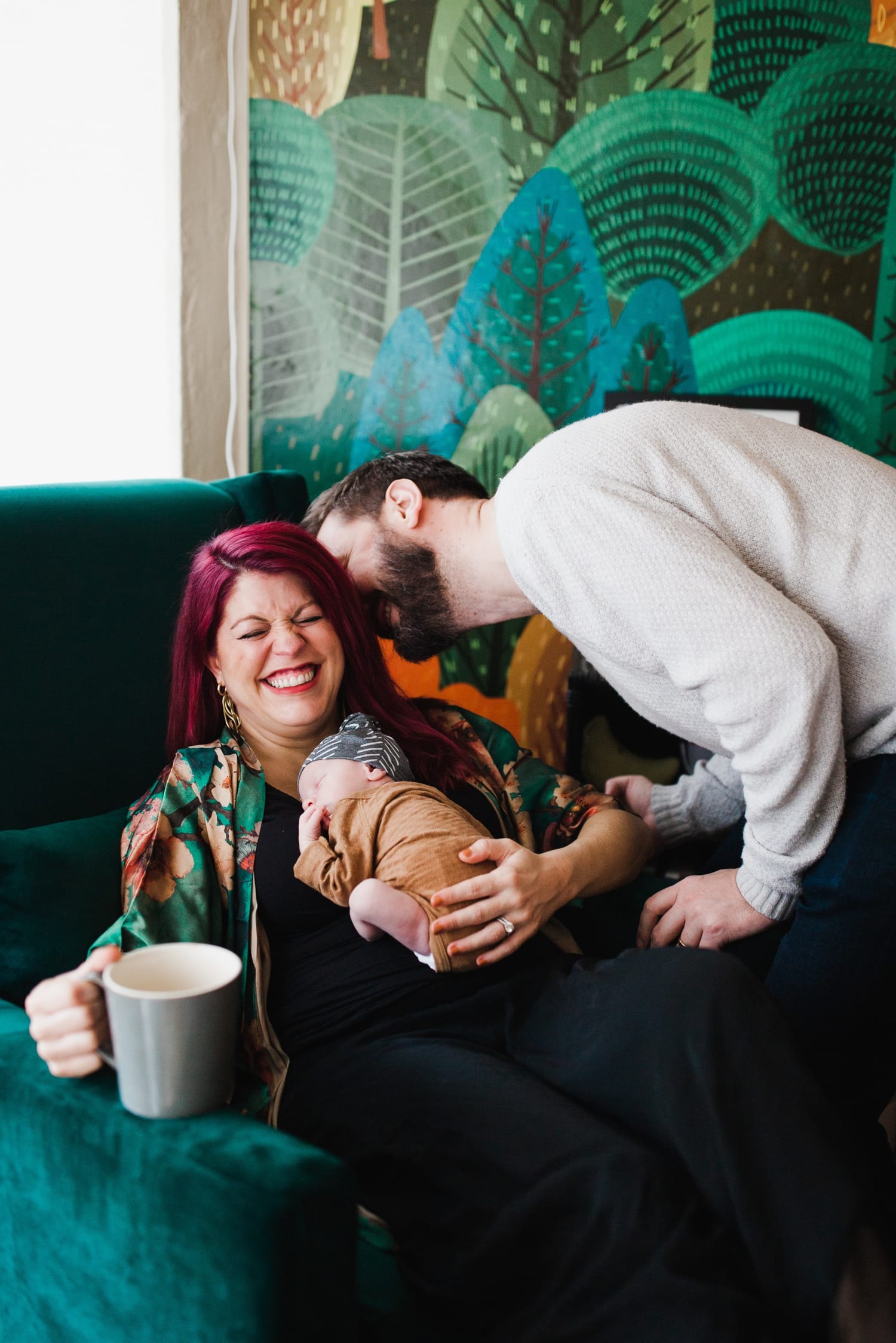 Mom laughs with her coffee as dad leans in over their sleeping newborn