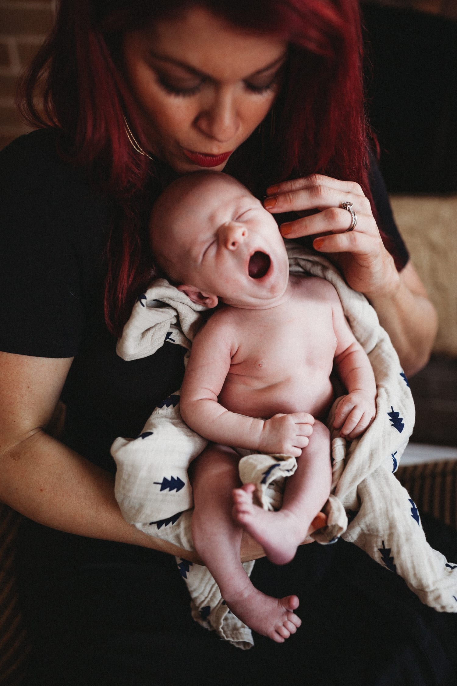 Newborn yawns wide in mom's arms, wrapped in a tree-print swaddle