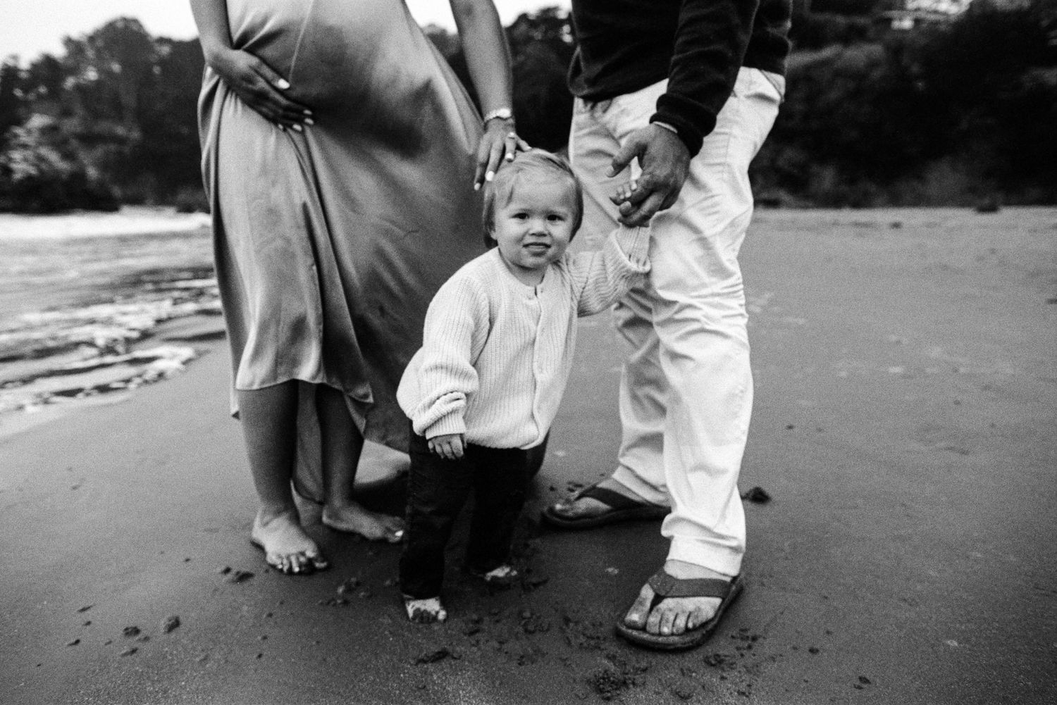 Black and white — toddler standing between parents at the beach during a maternity session