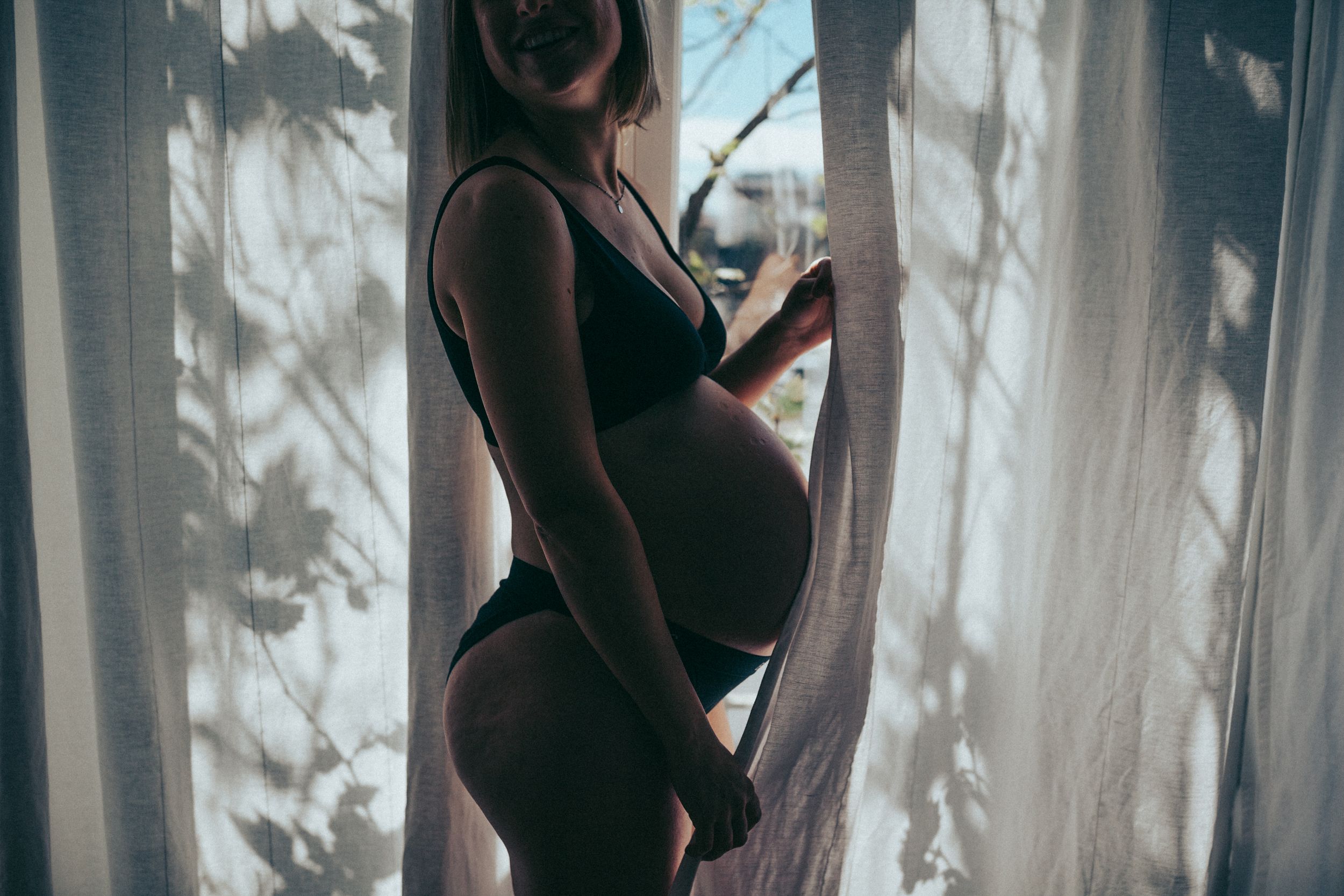 Silhouette of pregnant figure against sunlit curtains — in-home maternity photography
