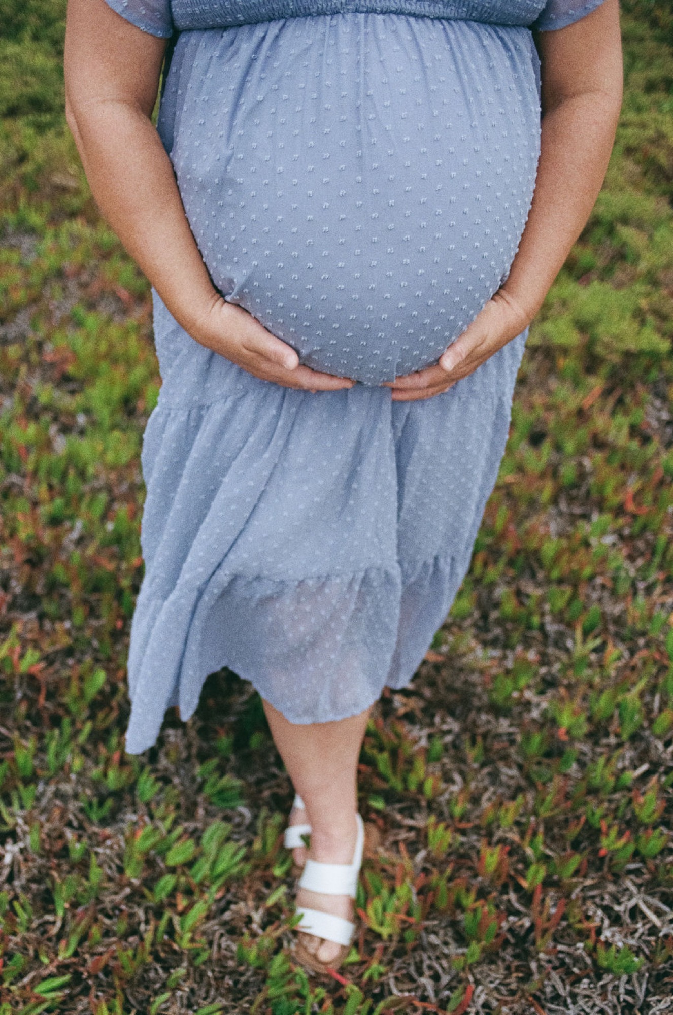 Pregnant woman cradling her belly in a blue dress over grassy terrain, hands gently framing the rounded midsection