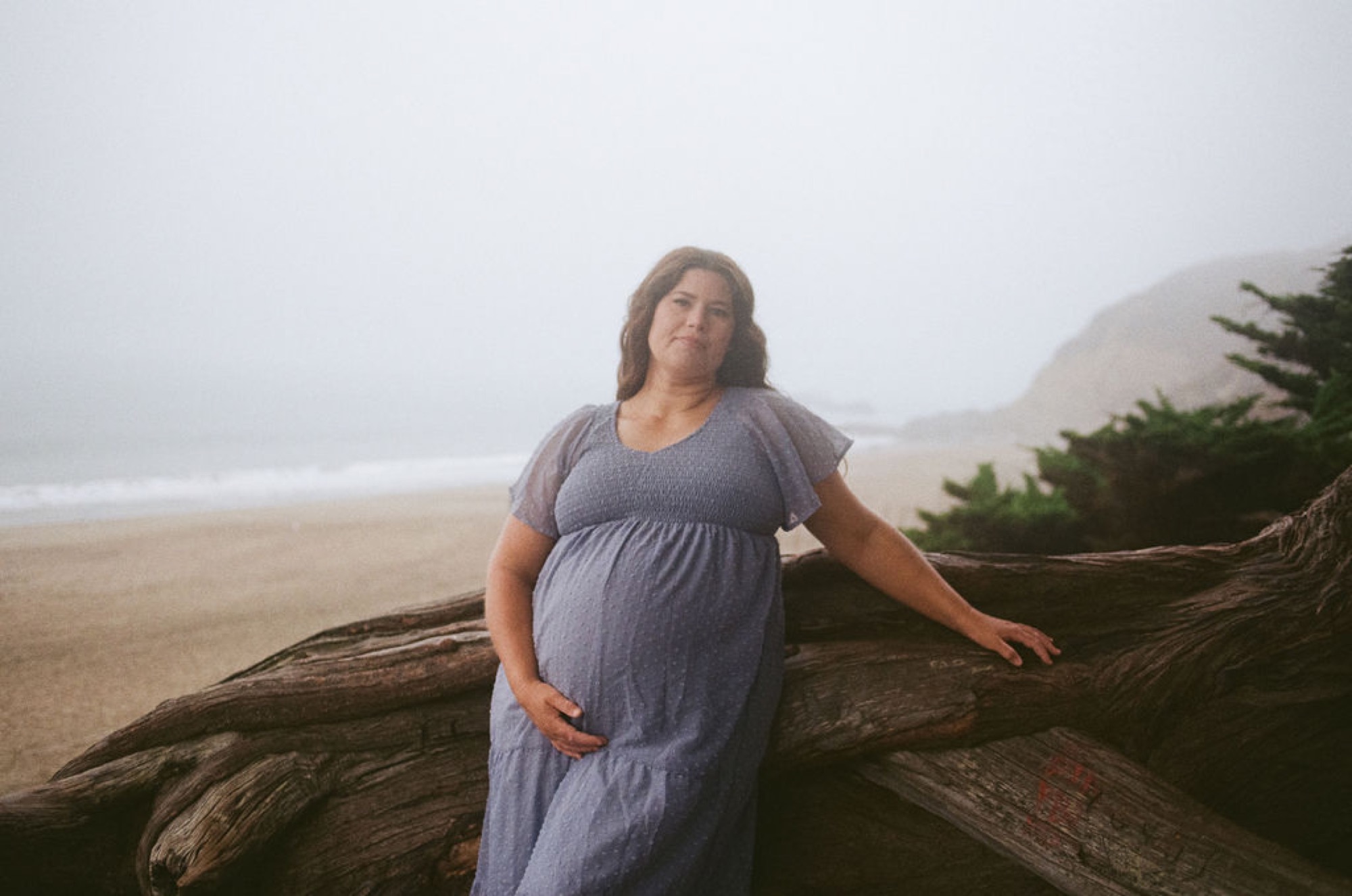 Pregnant woman in lavender dress rests against weathered driftwood on misty Baker Beach, mountains shrouded in fog behind her
