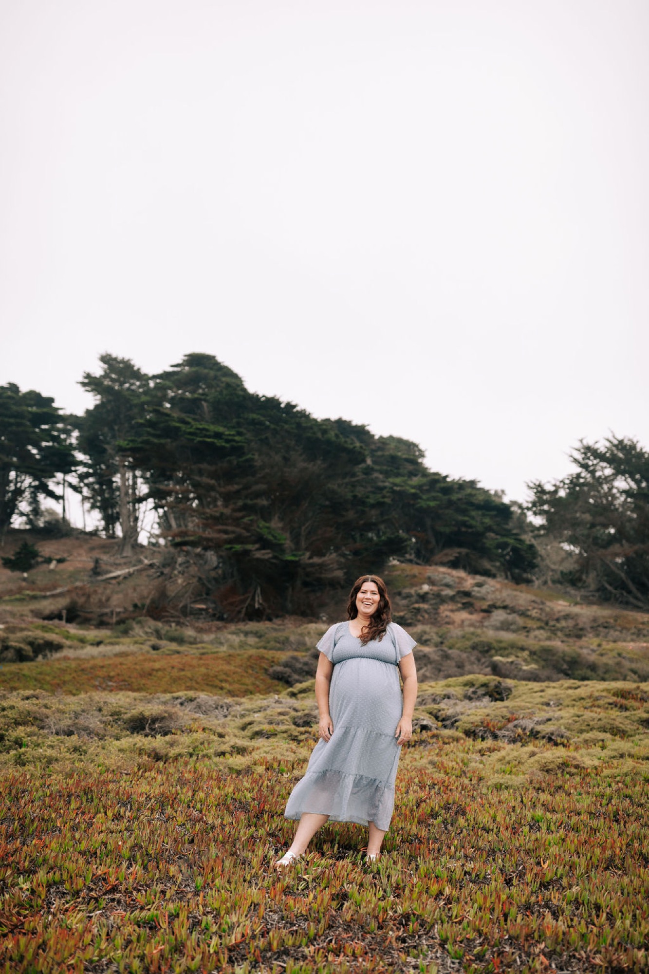 Expectant mother in a flowing gray dress stands among wildflowers on a windswept coastal hillside beneath overcast skies