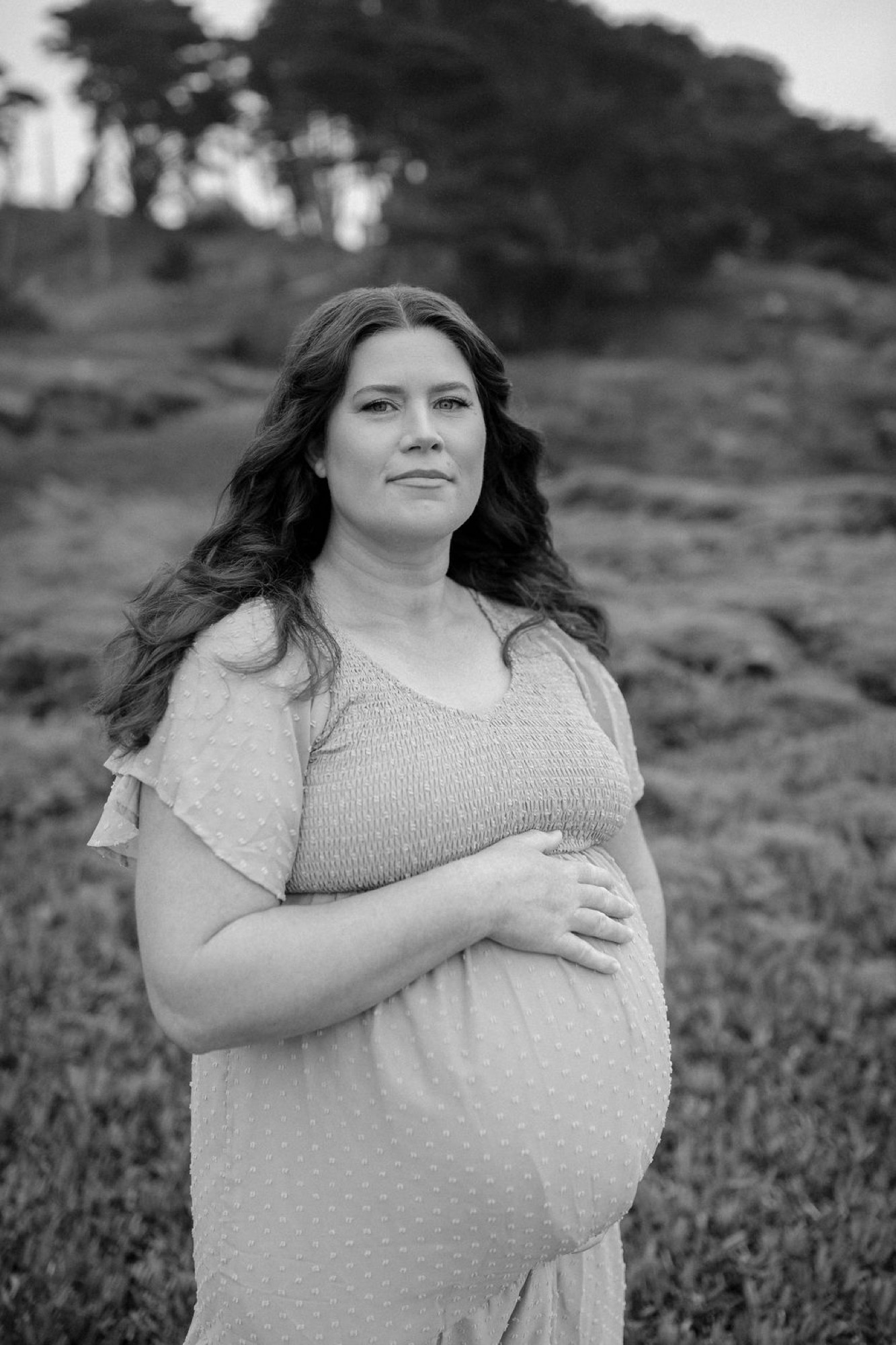 Pregnant woman in a fitted dress stands in an open grassy field, gazing upward with one hand cradling her belly against blurred hillside trees