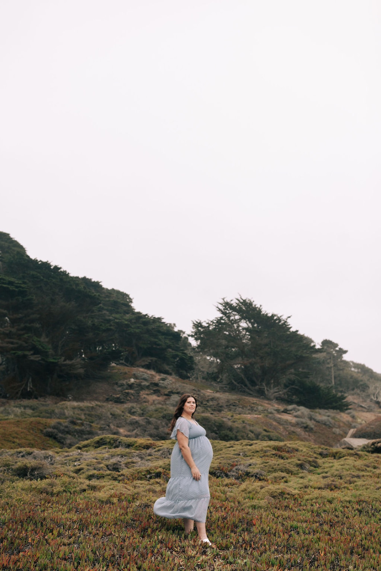Pregnant woman in white dress stands alone on windswept coastal hillside with dark cypress trees and overcast sky behind her