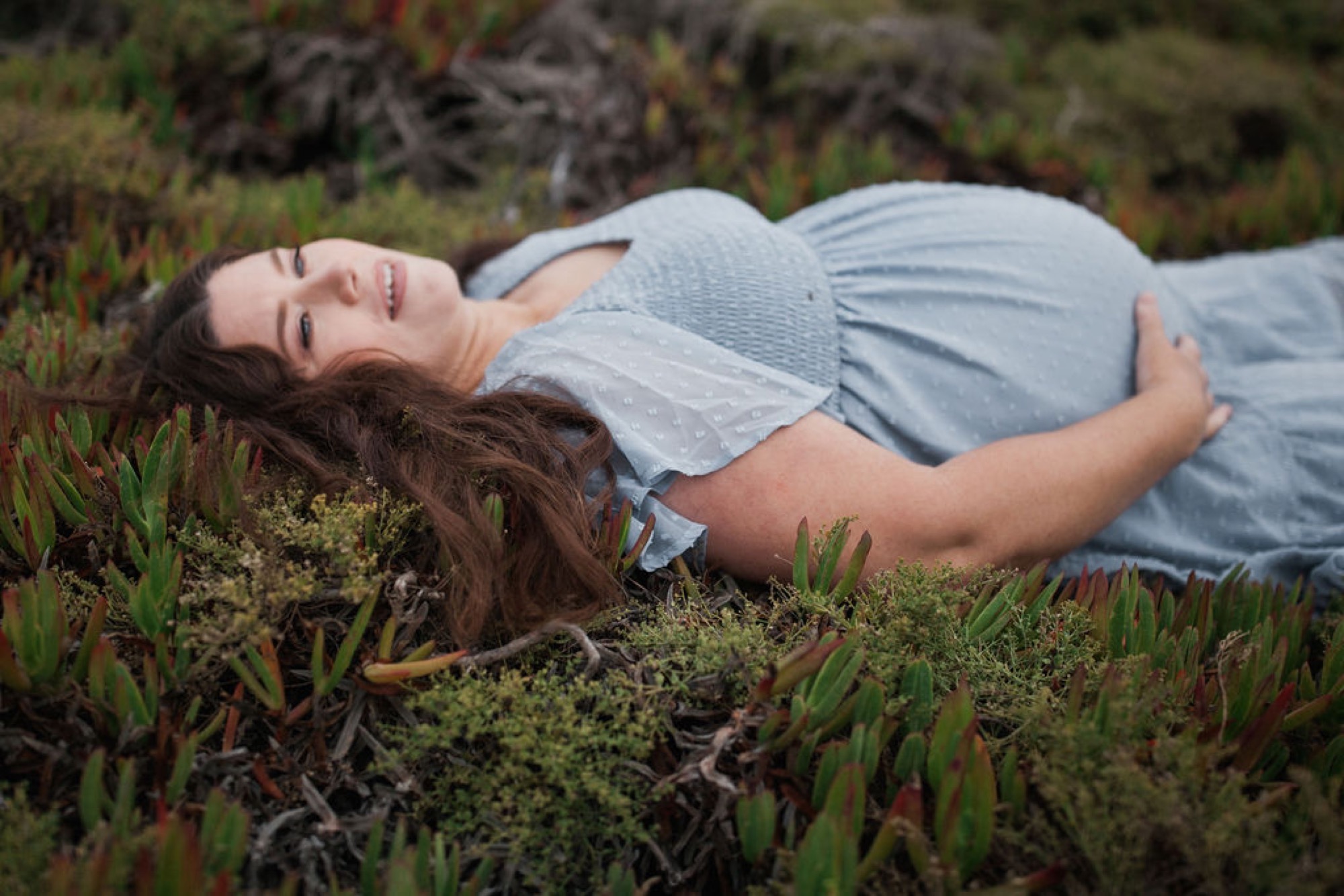Pregnant woman in light blue dress reclines on coastal moss and low vegetation, dark hair splayed across green plants, gazing upward with serene expression