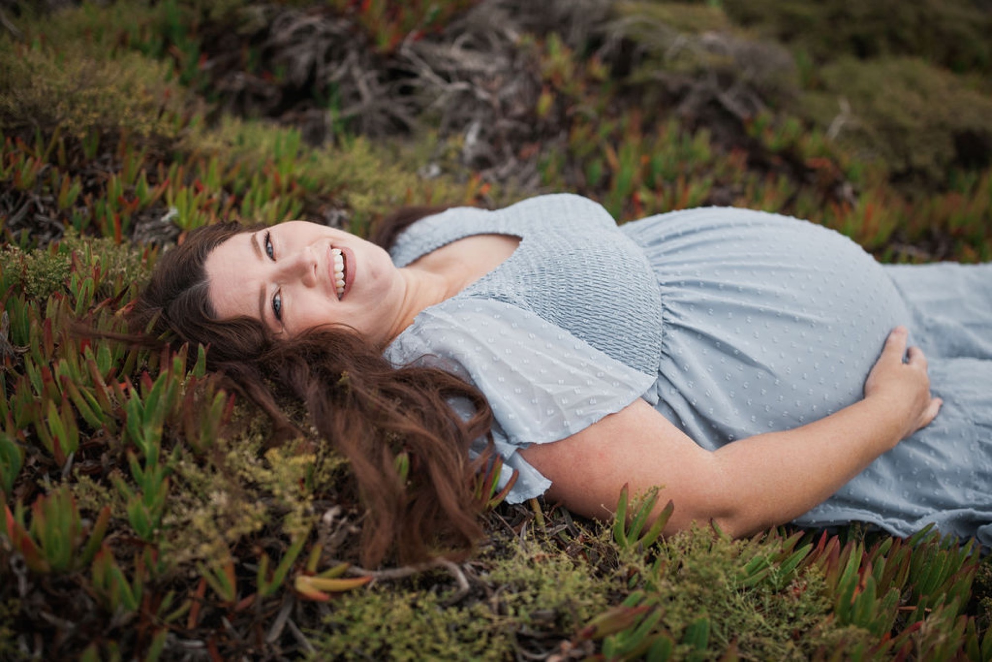 Pregnant woman lies contentedly on coastal scrub vegetation, cradling her belly with a genuine smile, her long dark hair cascading across the ground