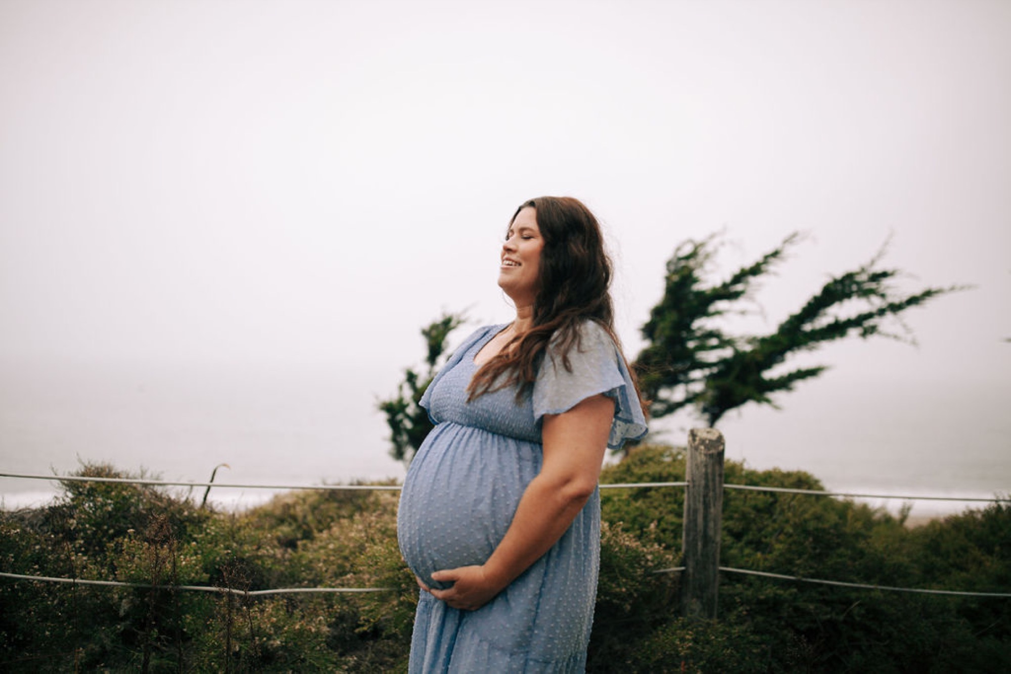 Pregnant woman in flowing blue dress gazes toward the horizon while cradling her belly, windswept coastal grasses and fence behind her