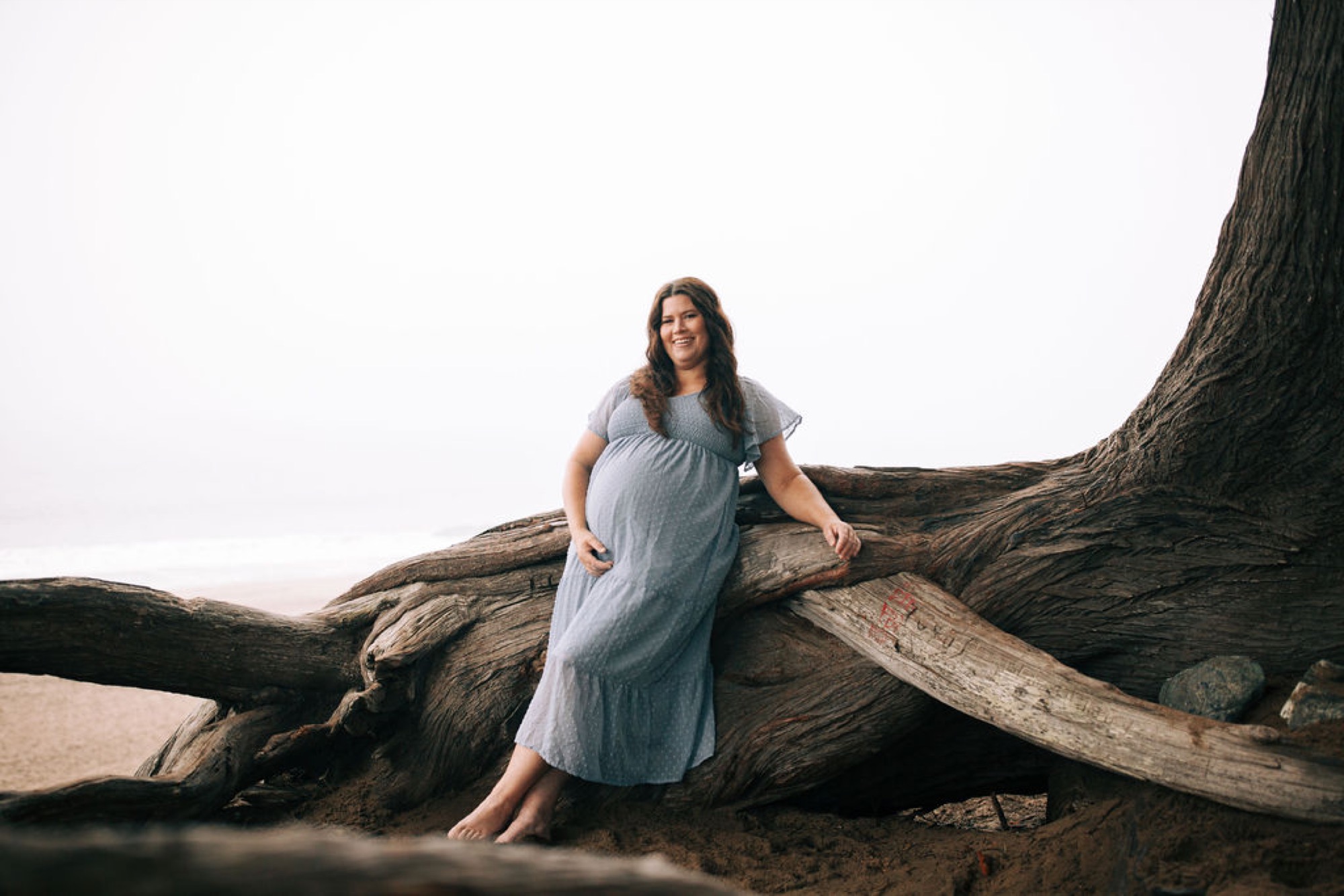 Pregnant woman in flowing blue dress leans against weathered driftwood on misty beach, gazing downward with serene expression