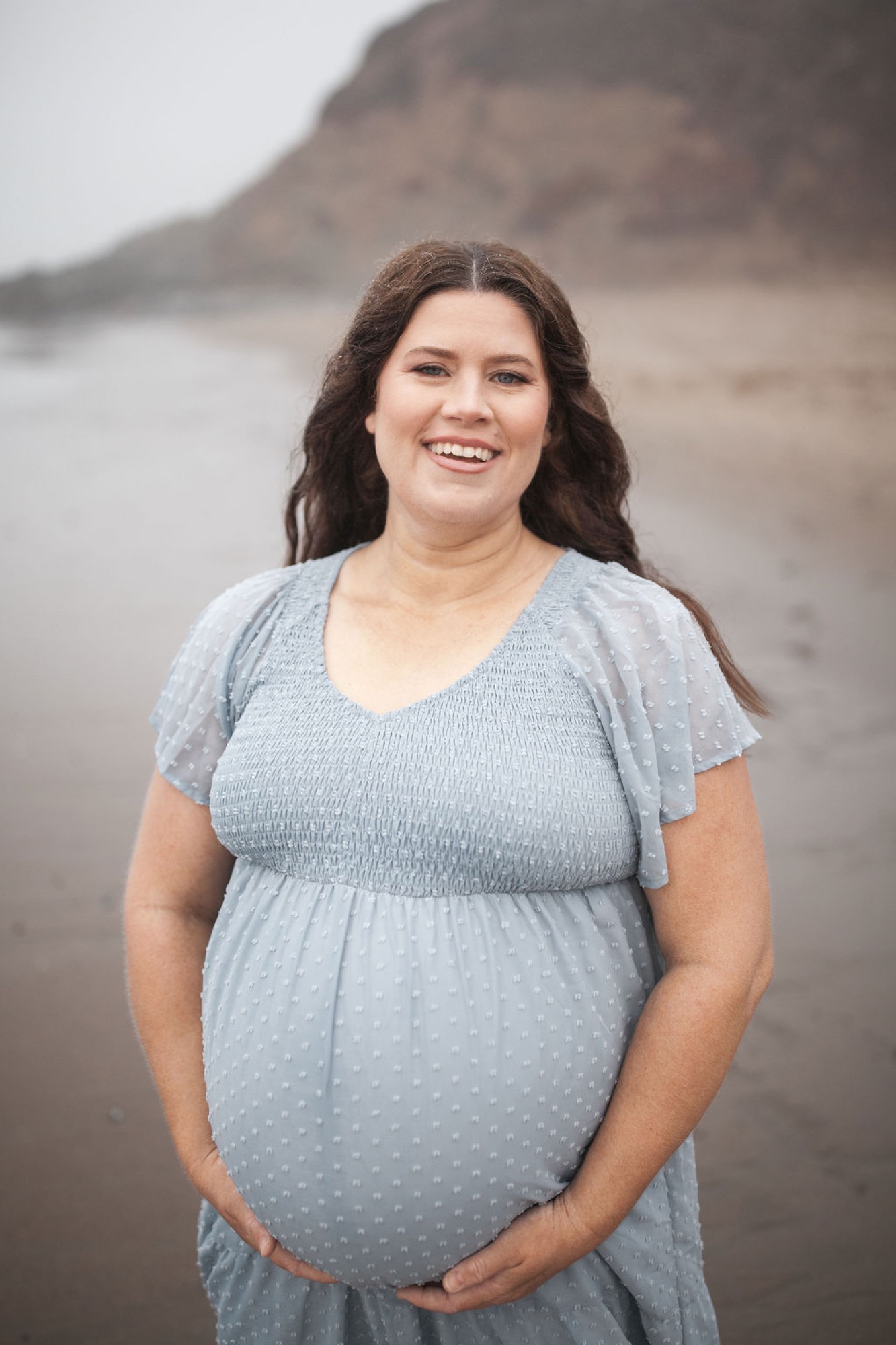 Pregnant woman in light blue maternity dress smiles warmly at camera with hand cradling belly, misty beach cliffs backdrop