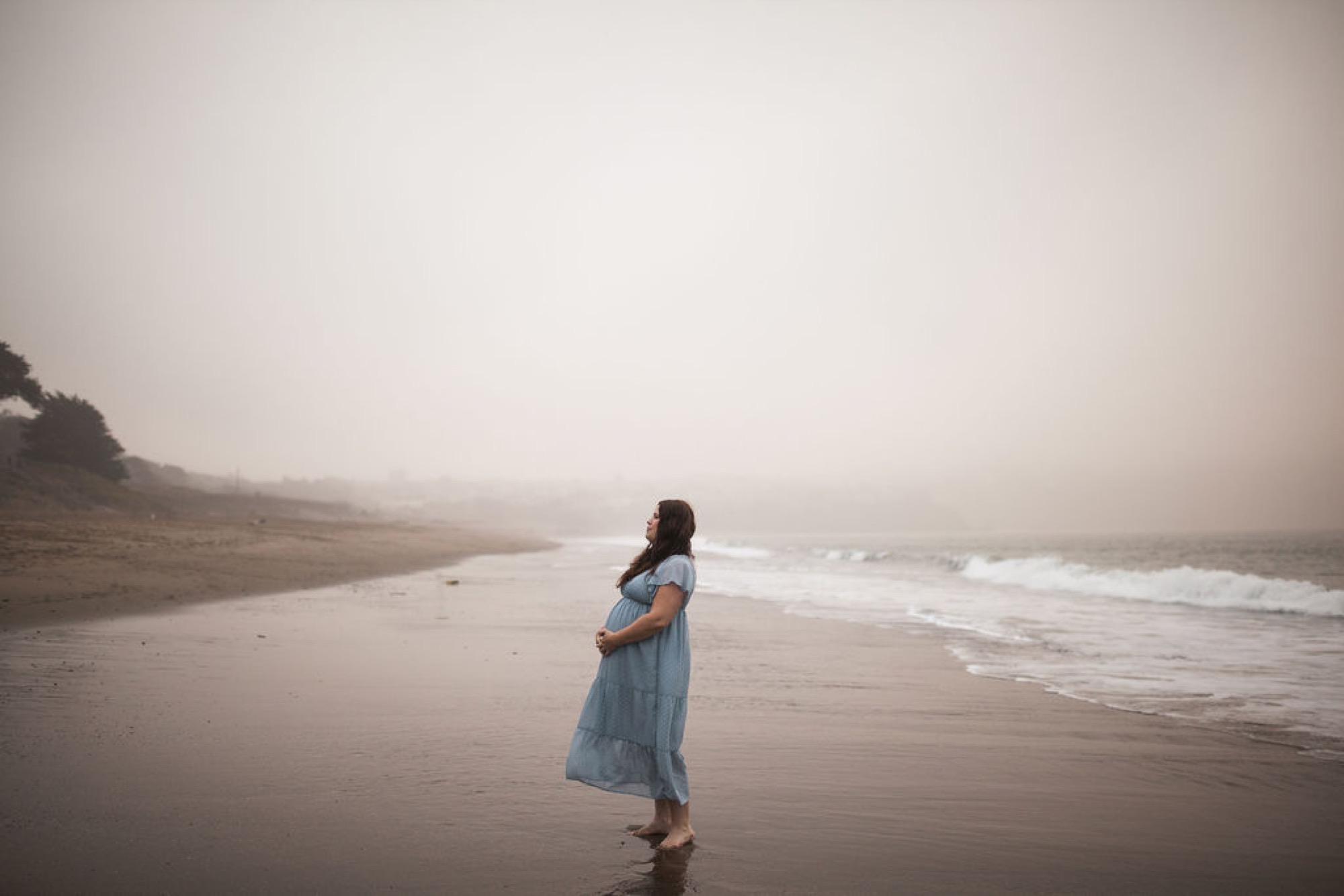Pregnant woman in a flowing blue dress stands alone on misty Baker Beach, gazing toward the gray ocean with waves rolling behind her