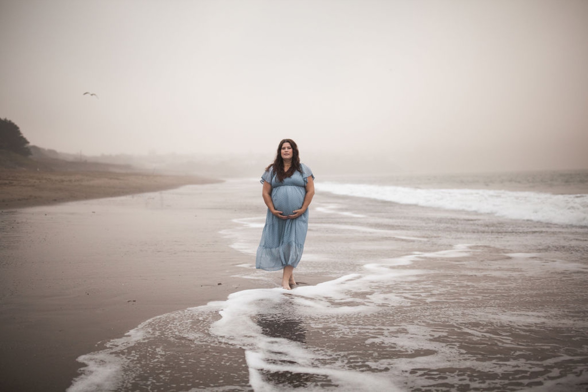 Pregnant woman in light blue dress stands barefoot in shallow ocean waves, gazing toward misty horizon under overcast coastal light
