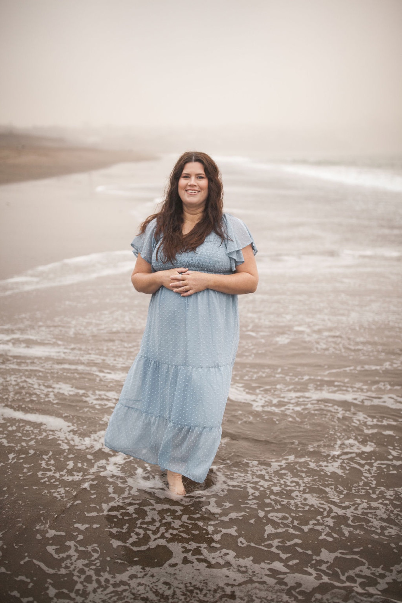Pregnant woman in light blue dress stands barefoot on wet sand, gazing toward camera with calm expression as waves lap around her feet
