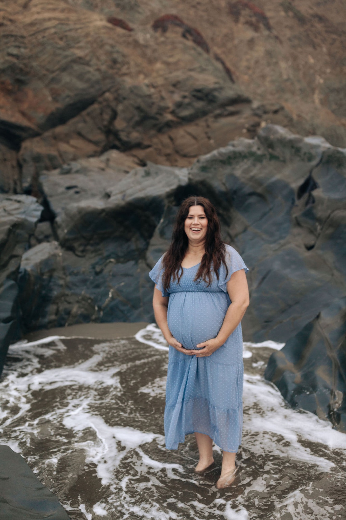Pregnant woman in light blue dress cradles her belly while standing barefoot in ocean waves against dramatic rocky cliffs