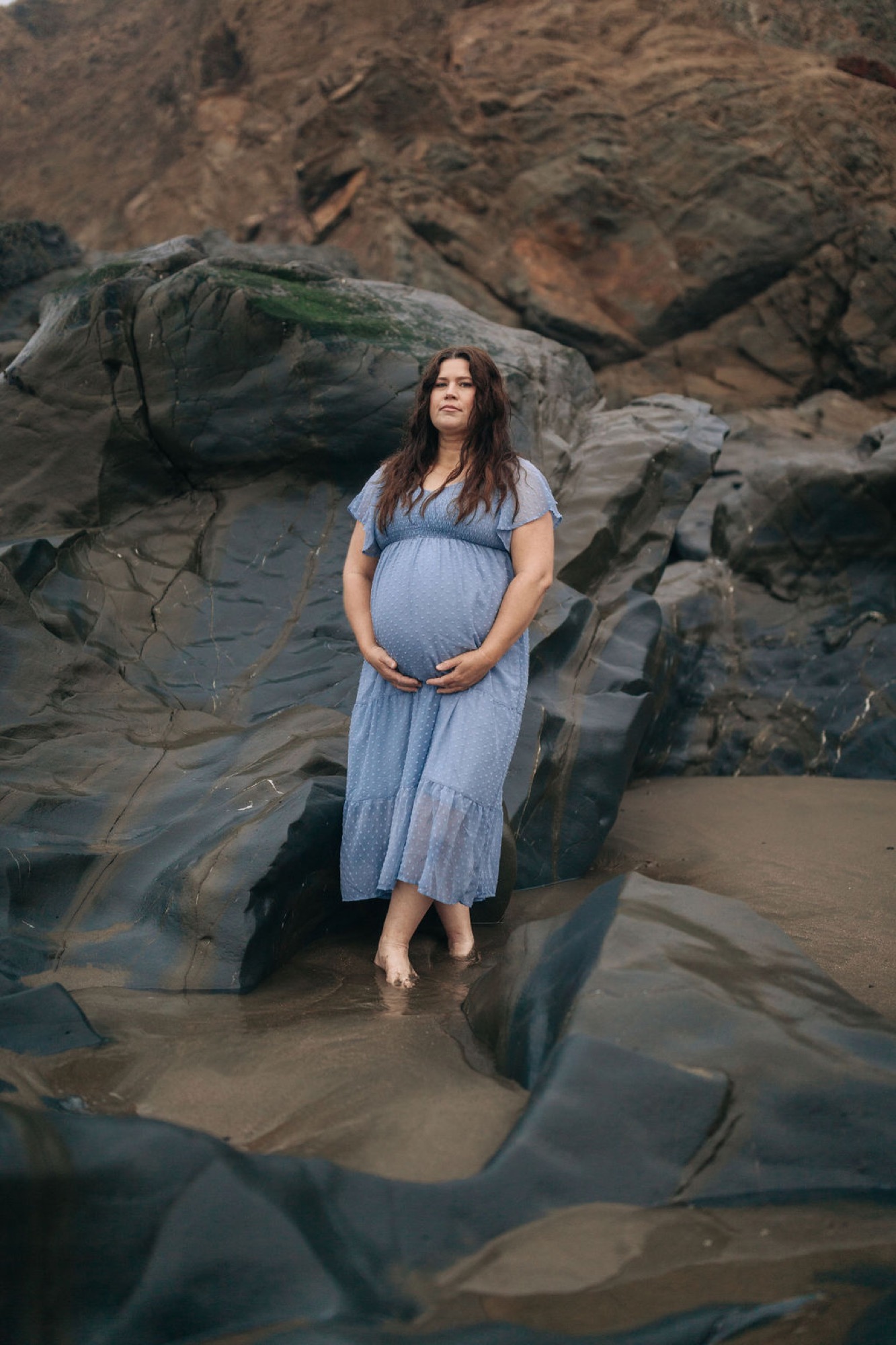 Pregnant woman in flowing blue dress stands contemplatively among weathered rocks, cradling her belly with serene expression