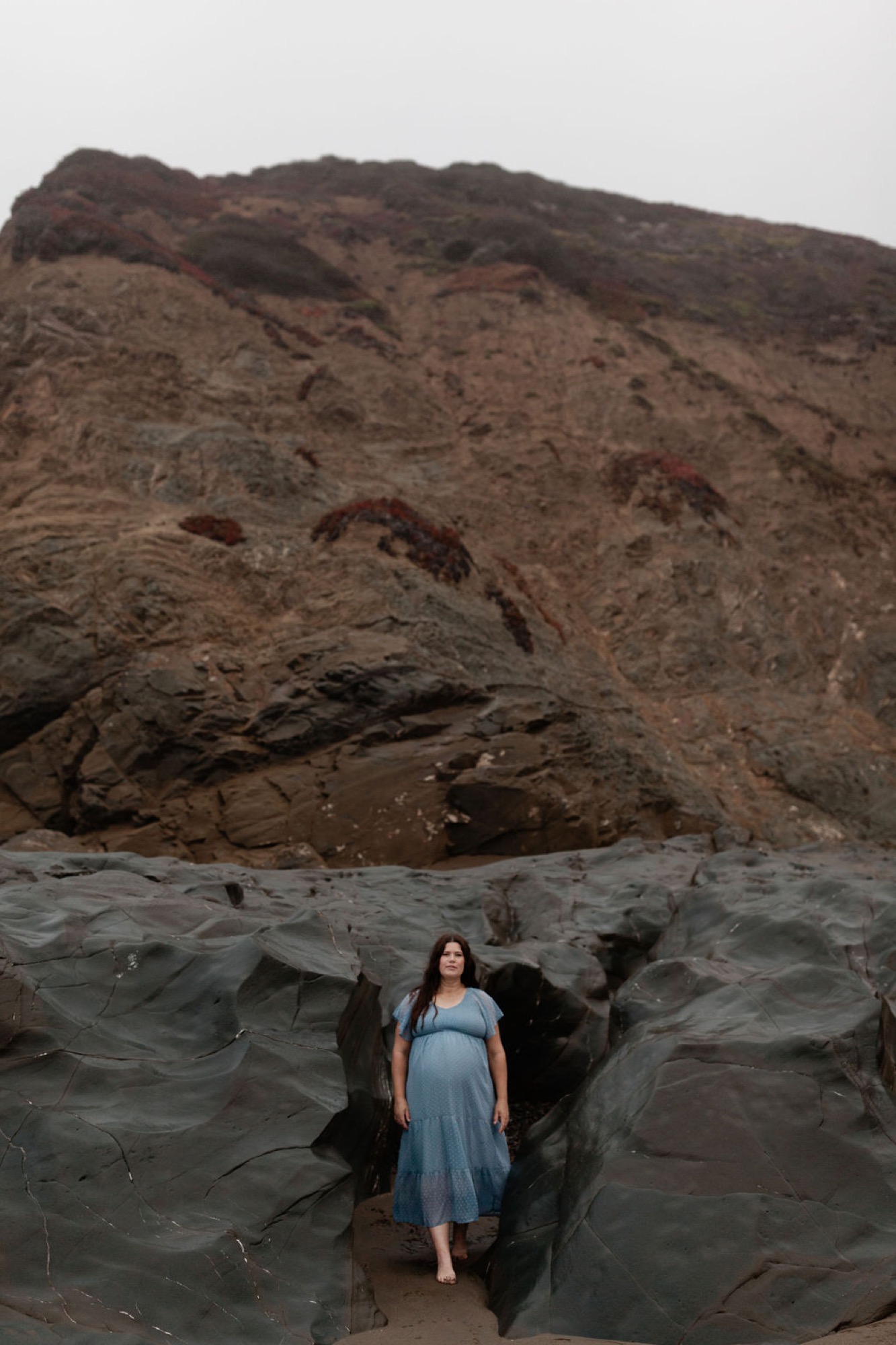 Pregnant woman in light blue dress stands nestled within dark rocky crevice beneath towering cliff face