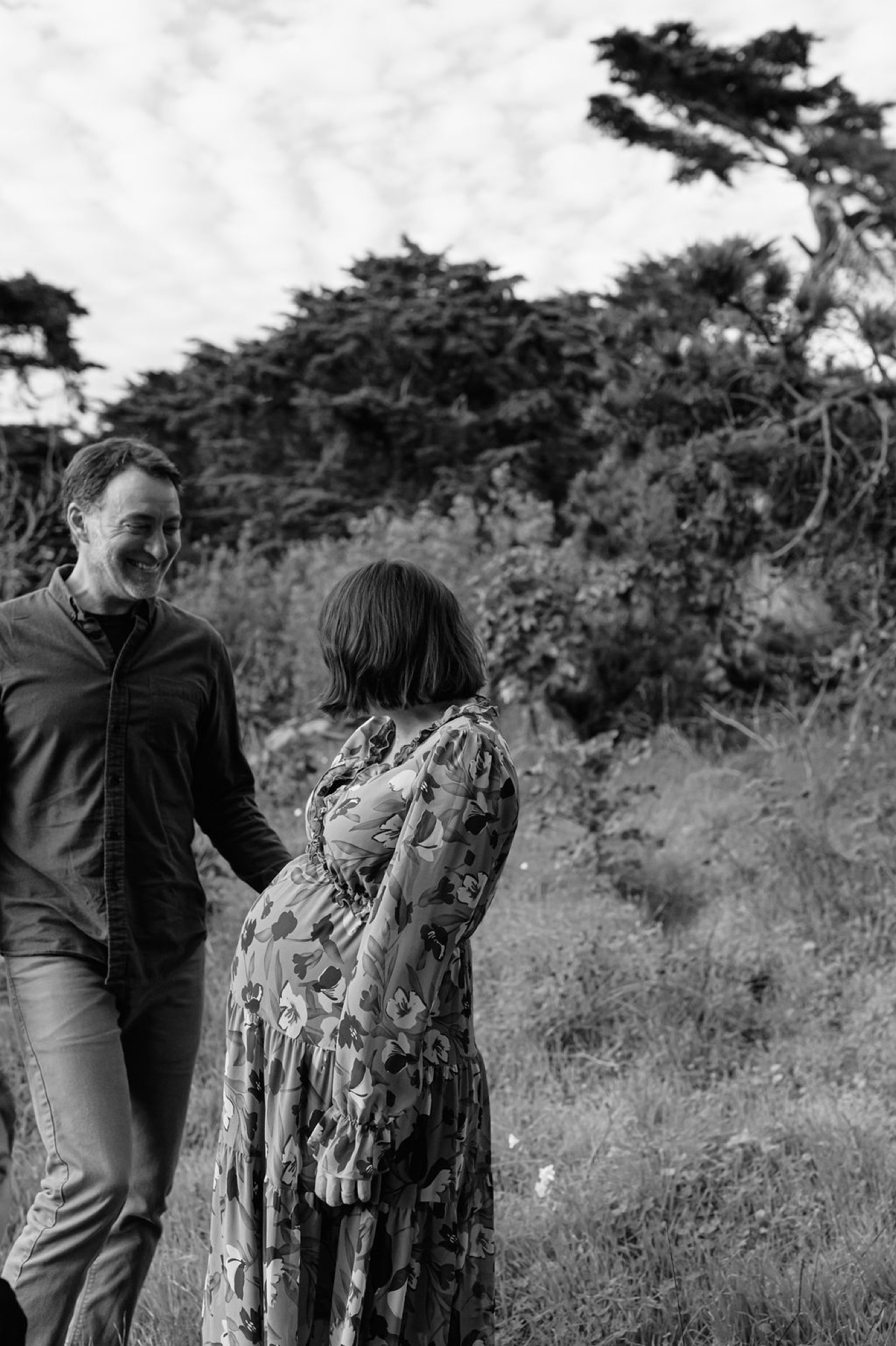 A pregnant woman in a floral dress walks beside her partner through windswept coastal grassland, her hand resting protectively on her belly
