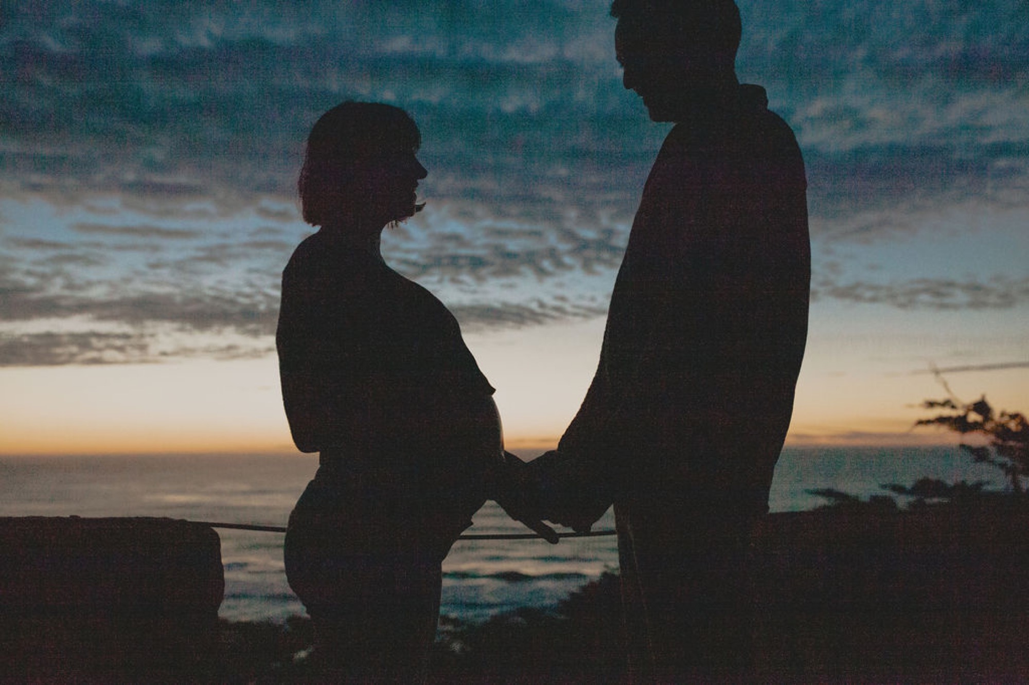 Silhouetted couple holds hands at sunset, the pregnant woman's profile glowing against the darkening ocean horizon