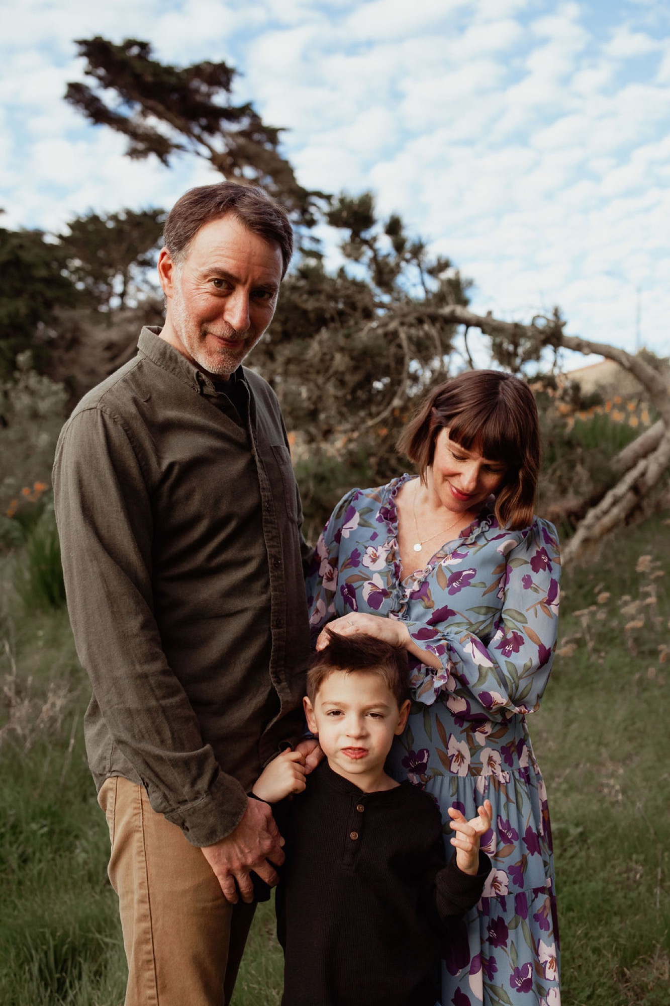 A family of three stands together on a grassy hillside with a windswept cypress tree behind them, the father and mother gazing warmly at their young child between them