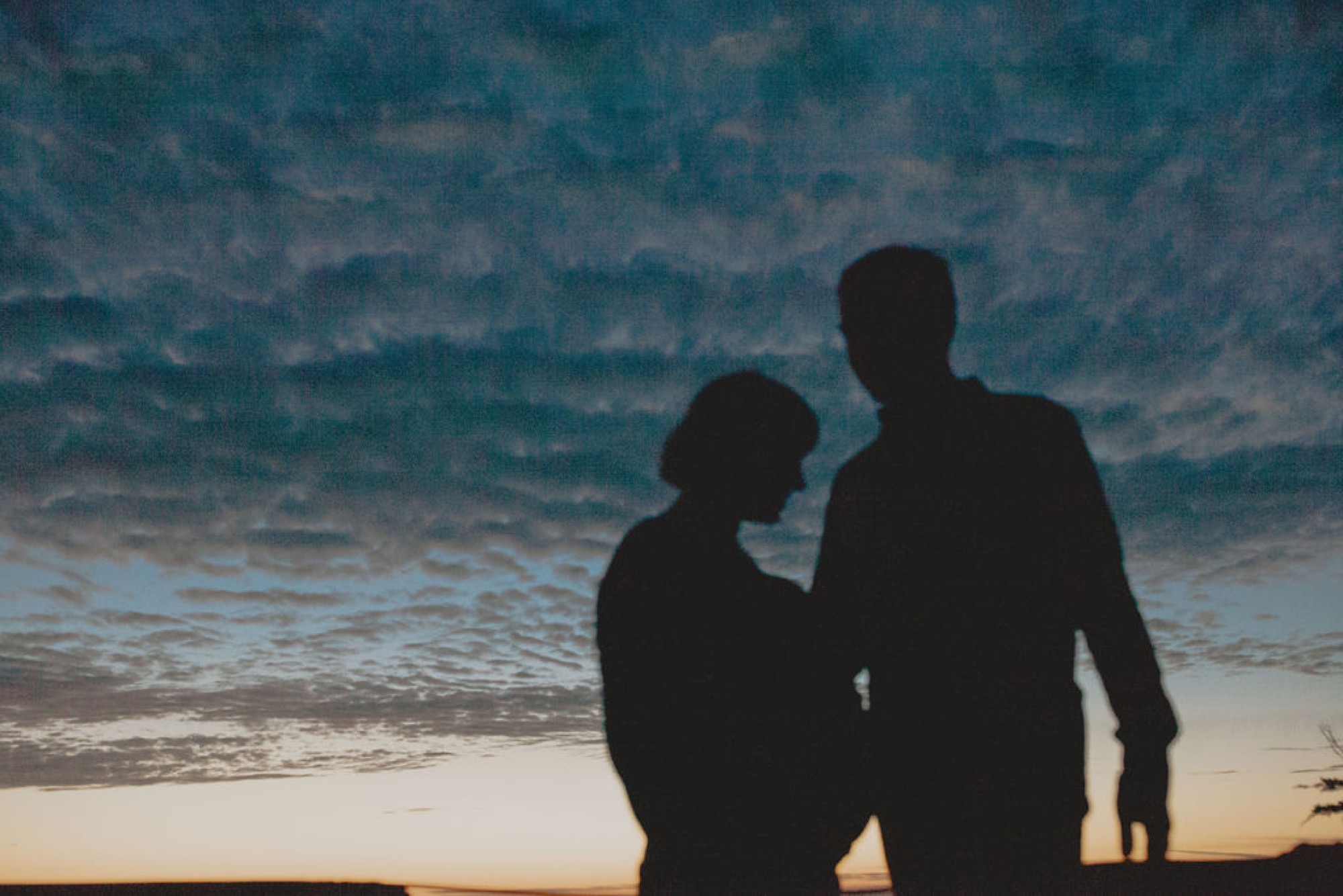 Two silhouetted figures stand close together against turbulent ocean waves and dramatic twilight sky, their profiles touching in intimate embrace
