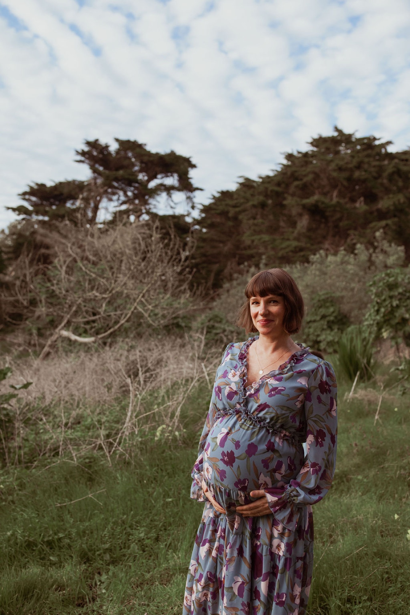 Pregnant woman in floral dress stands among windswept coastal trees, hand cradling belly, soft afternoon light filtering through branches