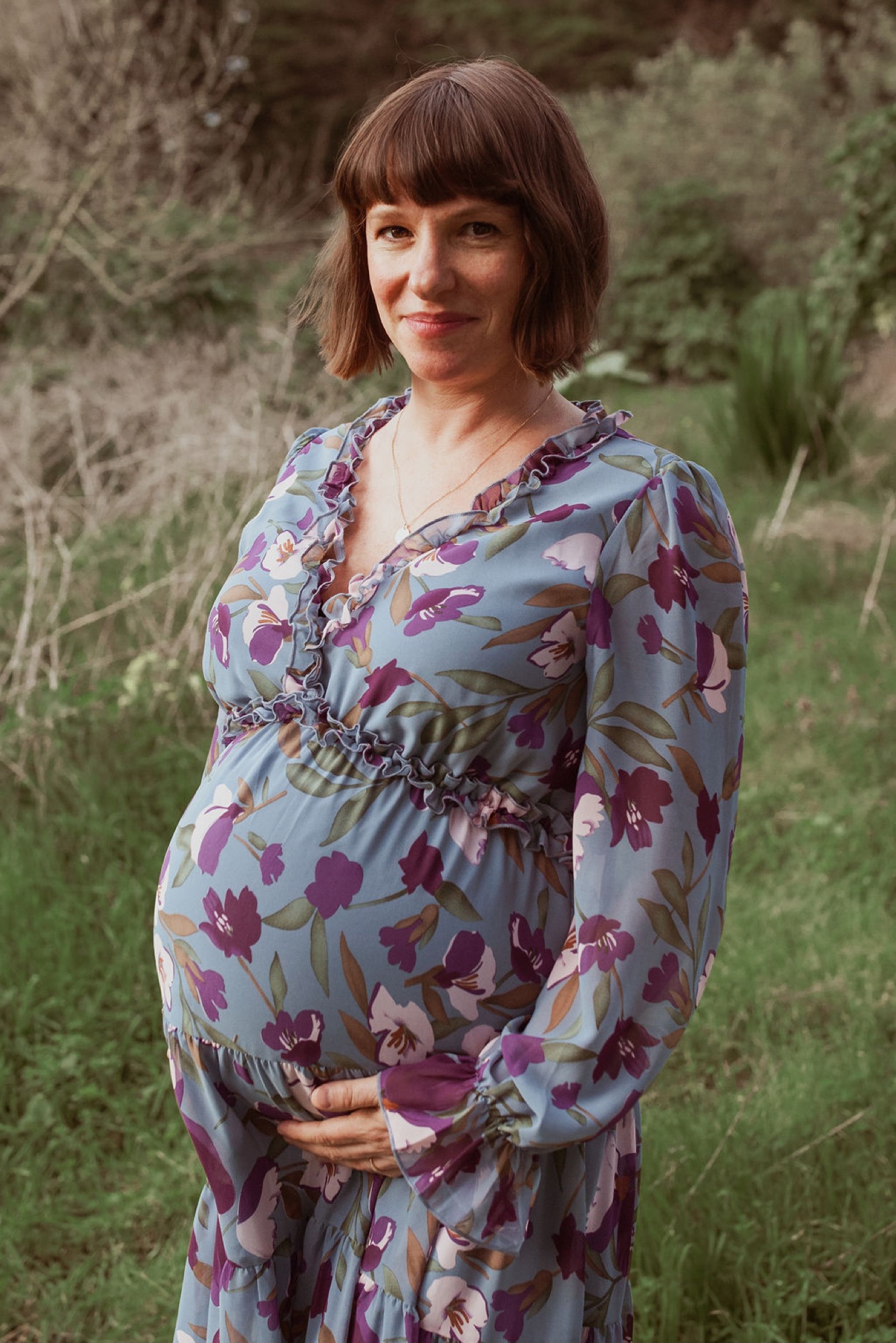 Pregnant woman in purple floral wrap dress cradling her belly while standing in a sunlit woodland clearing with soft natural light filtering through trees