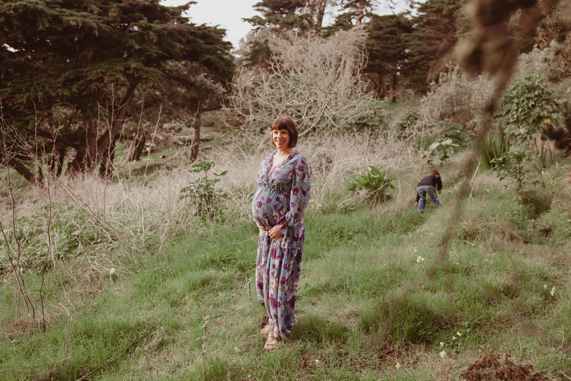 Pregnant woman in purple patterned dress stands among tall grasses and evergreen trees, bathed in soft natural light