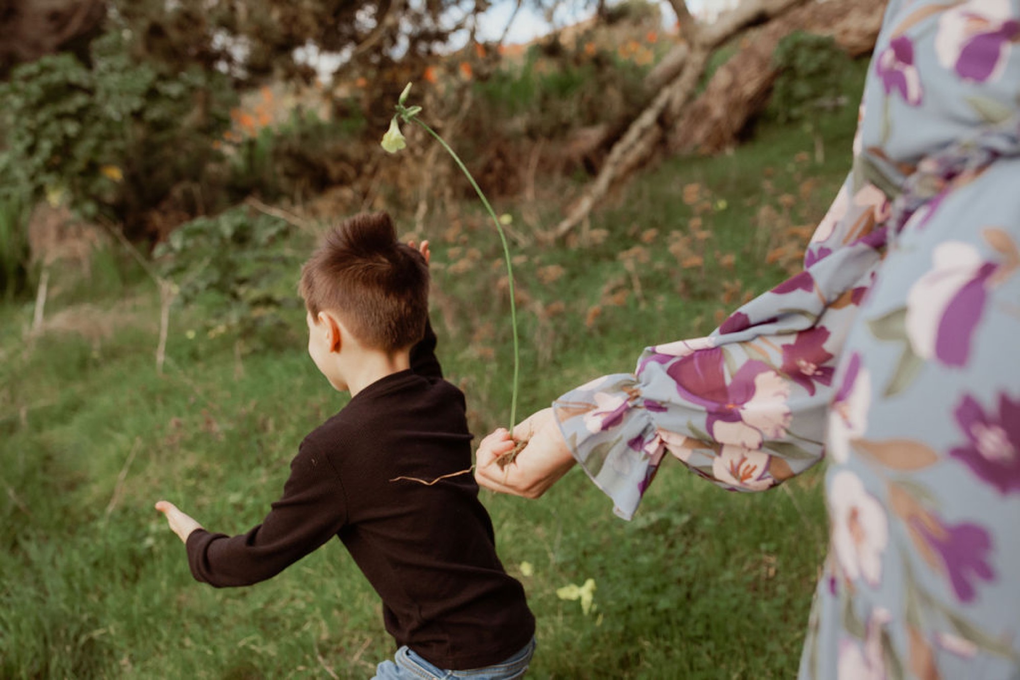 A child in a dark jacket reaches toward an adult's floral-patterned sleeve while holding a thin branch, engaged in gentle play on a grassy hillside