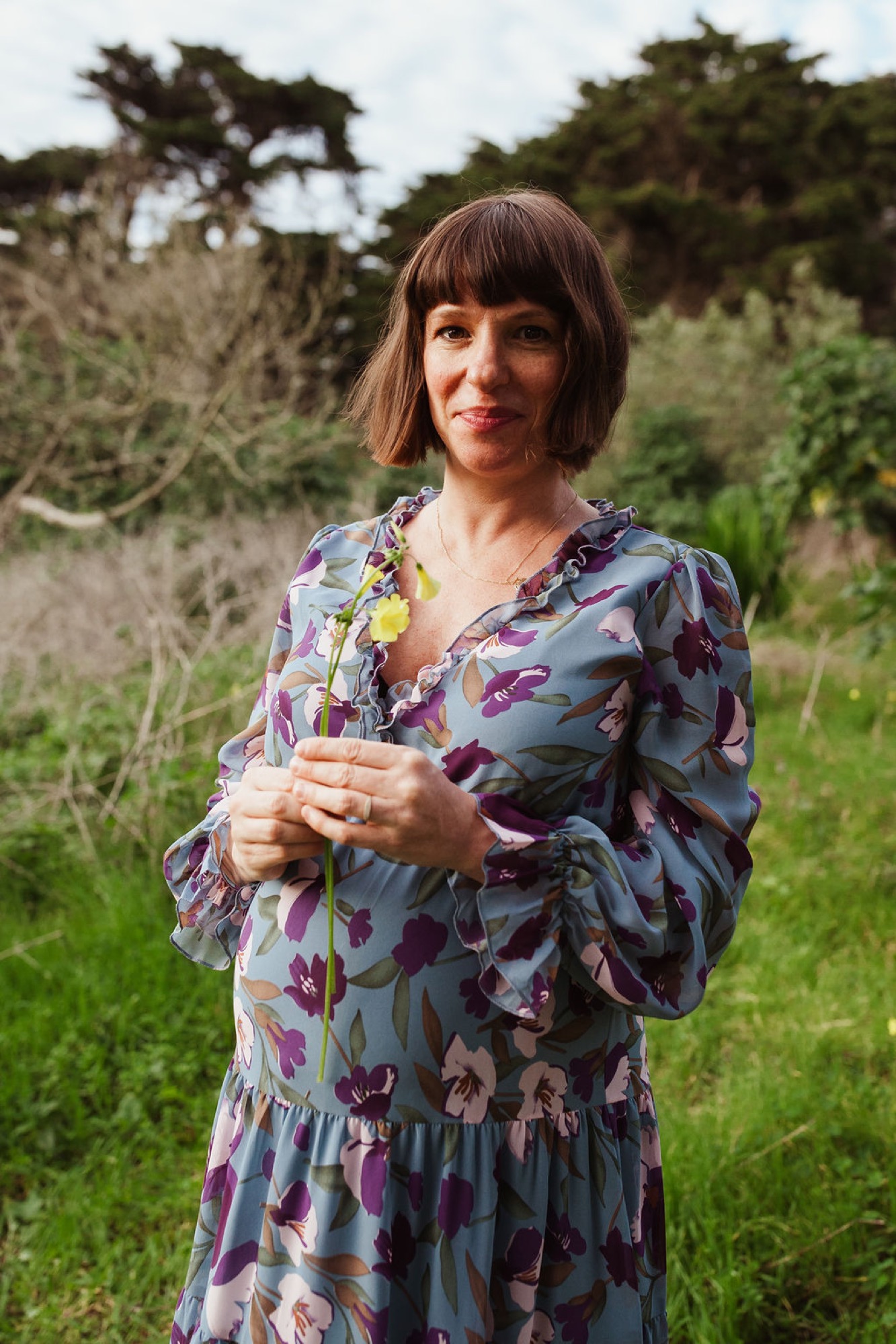 Expectant mother in a colorful floral wrap dress stands in a sunlit meadow with windswept cypress trees framing the natural landscape behind her