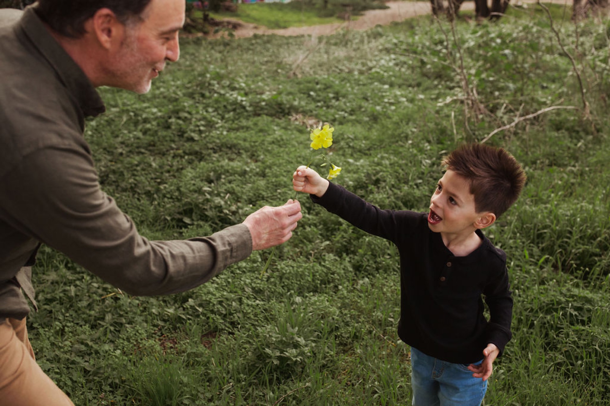 A man hands a yellow flower to a young child in a lush green garden setting, both reaching toward each other with genuine joy
