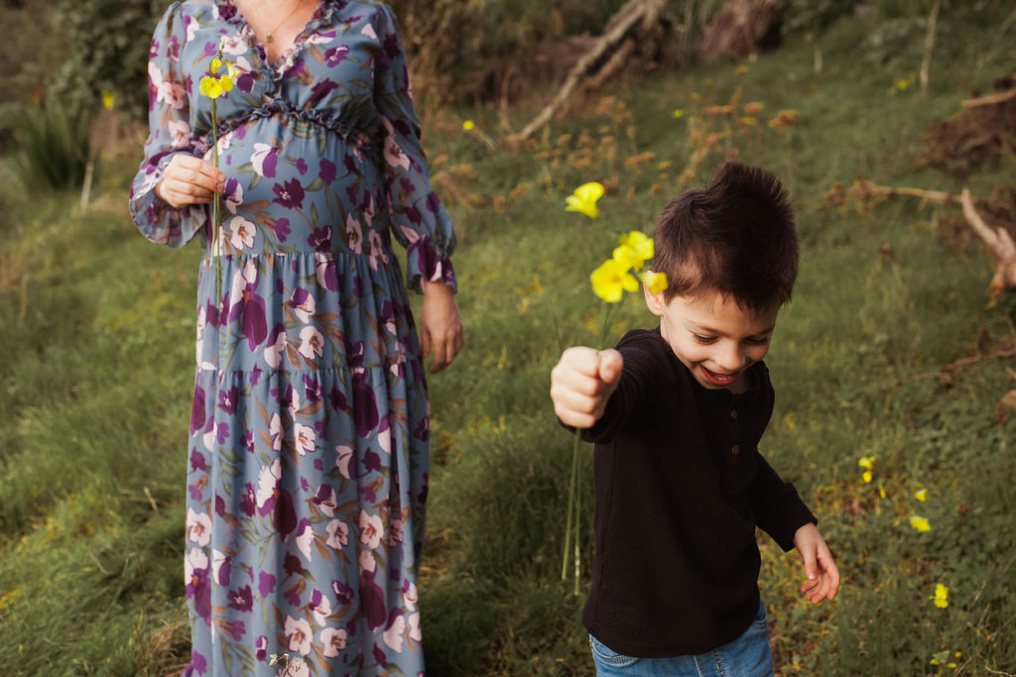 A young boy in a black hoodie reaches toward yellow wildflowers while standing beside a pregnant woman in a floral dress on a grassy hillside