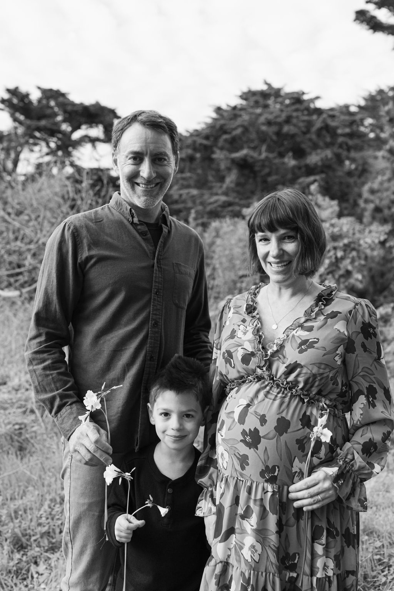 Family of three smiling at camera in natural meadow setting, woman visibly pregnant in floral dress, man in dark jacket with hand on child's shoulder
