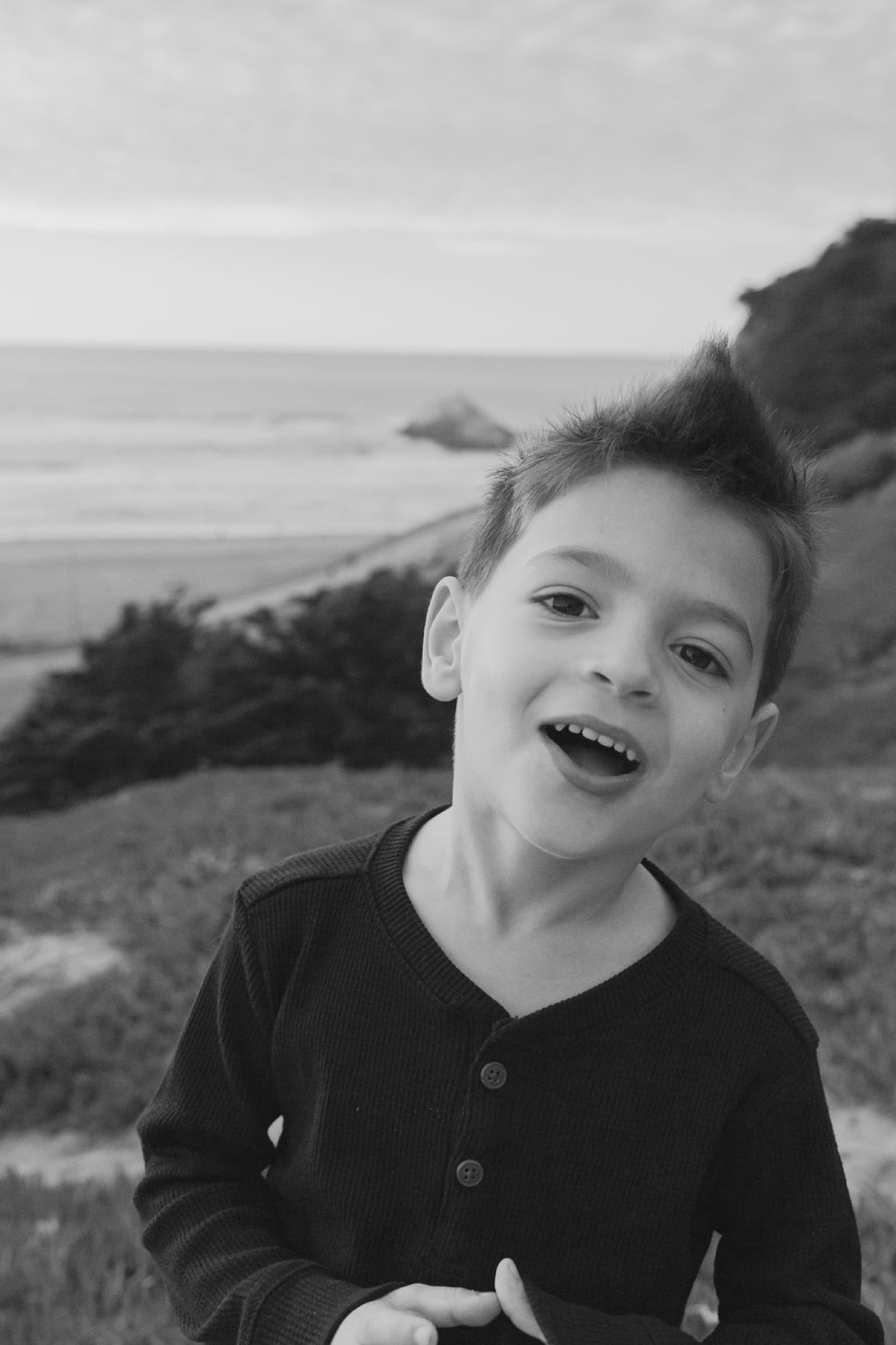 Young boy laughs joyfully at coastal overlook with bay waters and rocky cliffs stretching behind him in soft natural light