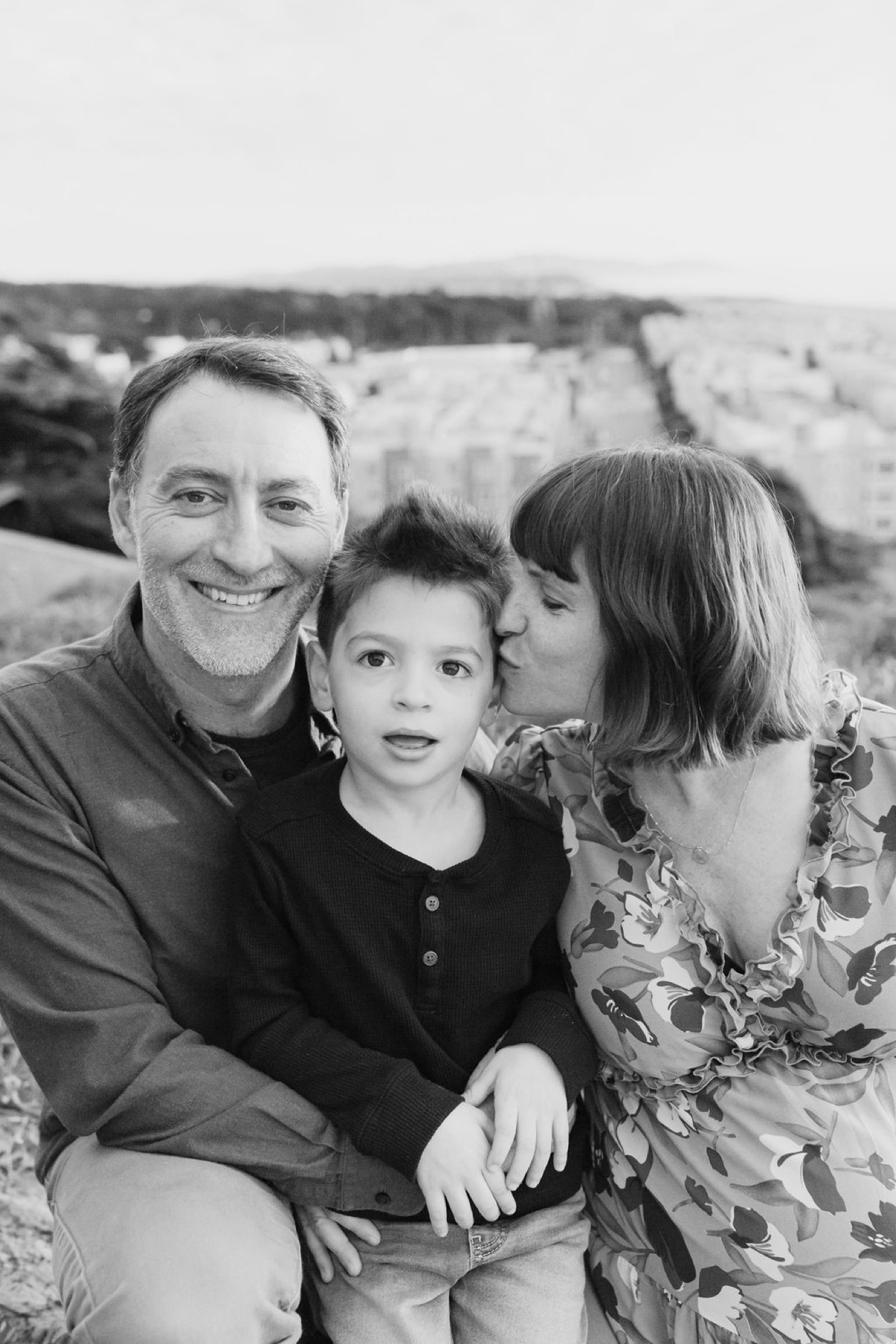 A man and woman embrace their young son between them on a windswept hillside, all three smiling broadly at the camera in soft natural light